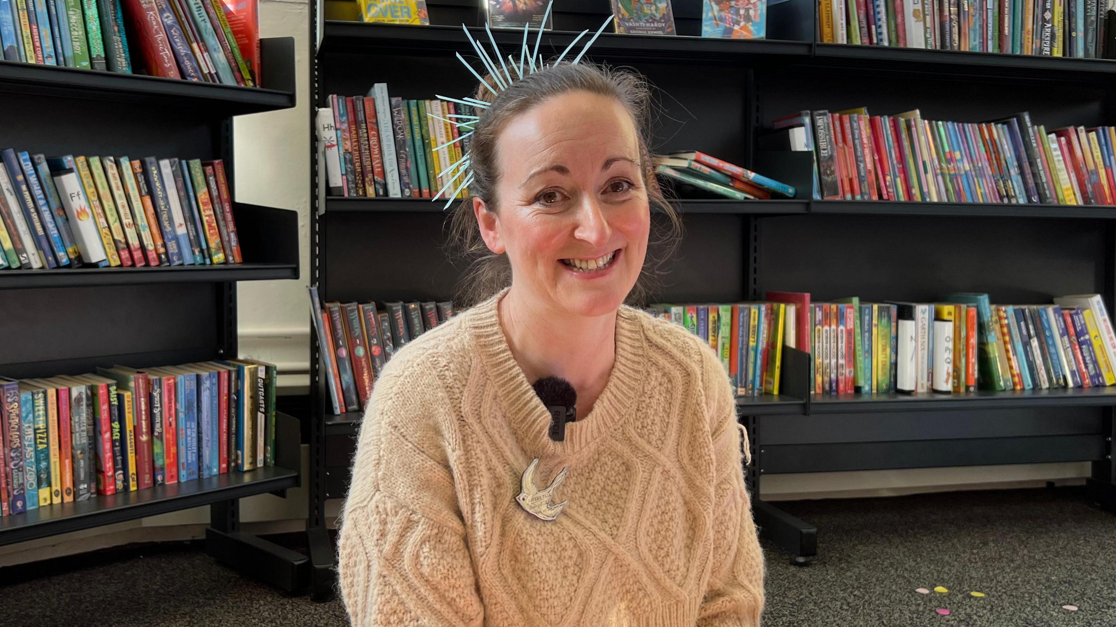 Joanna Grace sits in a library in front of two book shelves. She is smiling and has a spiky fascinator on the back of her head. She is wearing a cream-coloured jumper and has a bird badge on it.