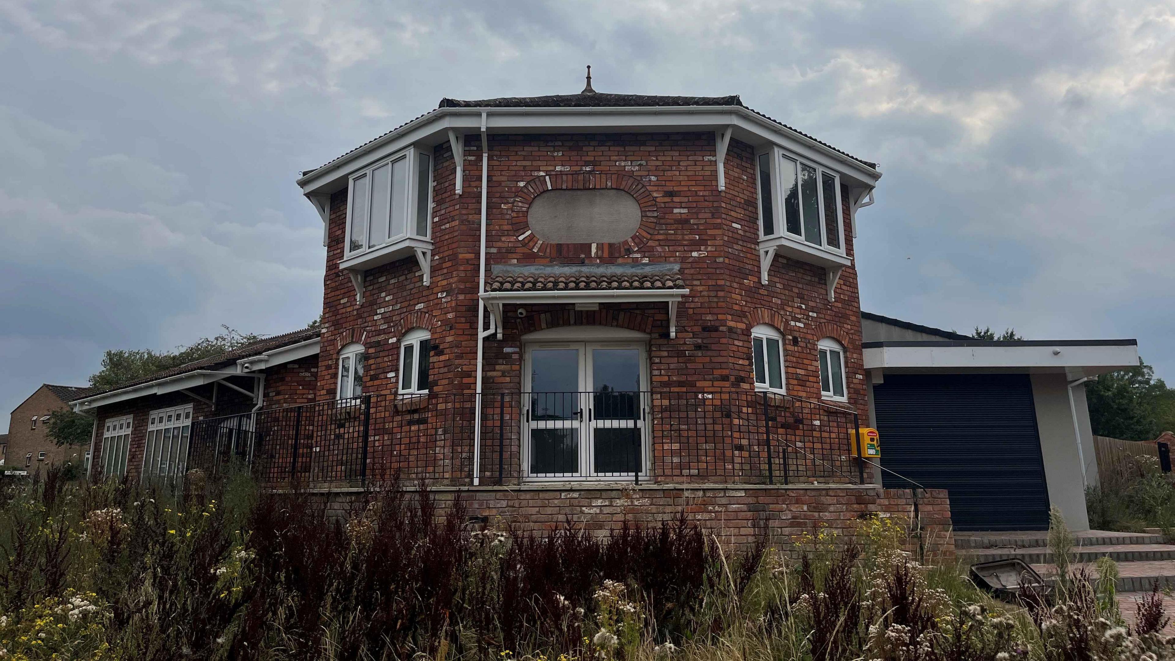 The exterior of a red building that has a hexagonal entrance. To the right there is an attached black garage. In the foreground is an overgrown garden area. 