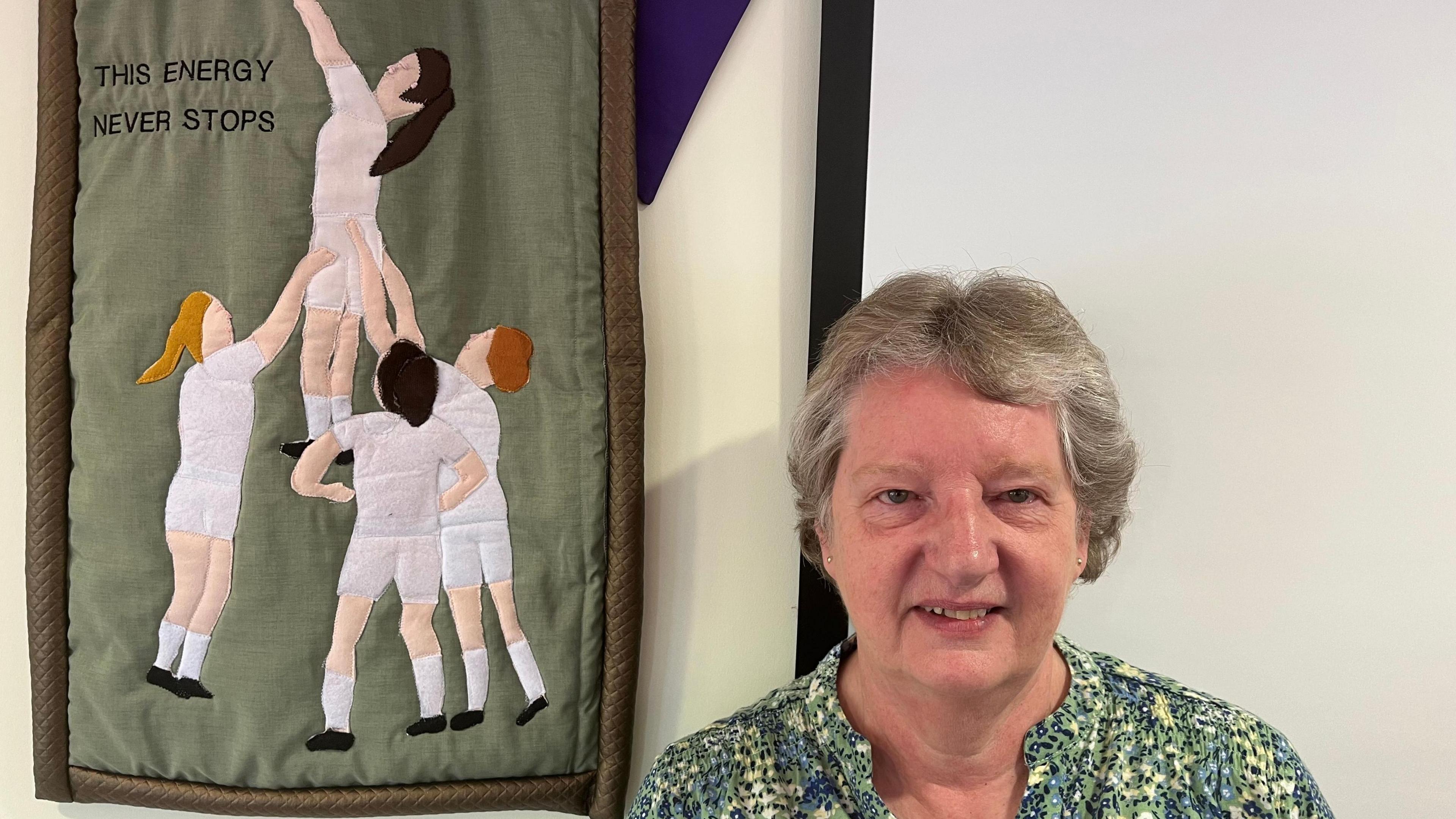 A knitted mural with three woman in white rugby kit holding up another woman in a line out. The words "This energy never stops" is knitted into the mural. In front of the mural, a woman in a green shirt looks at the camera.