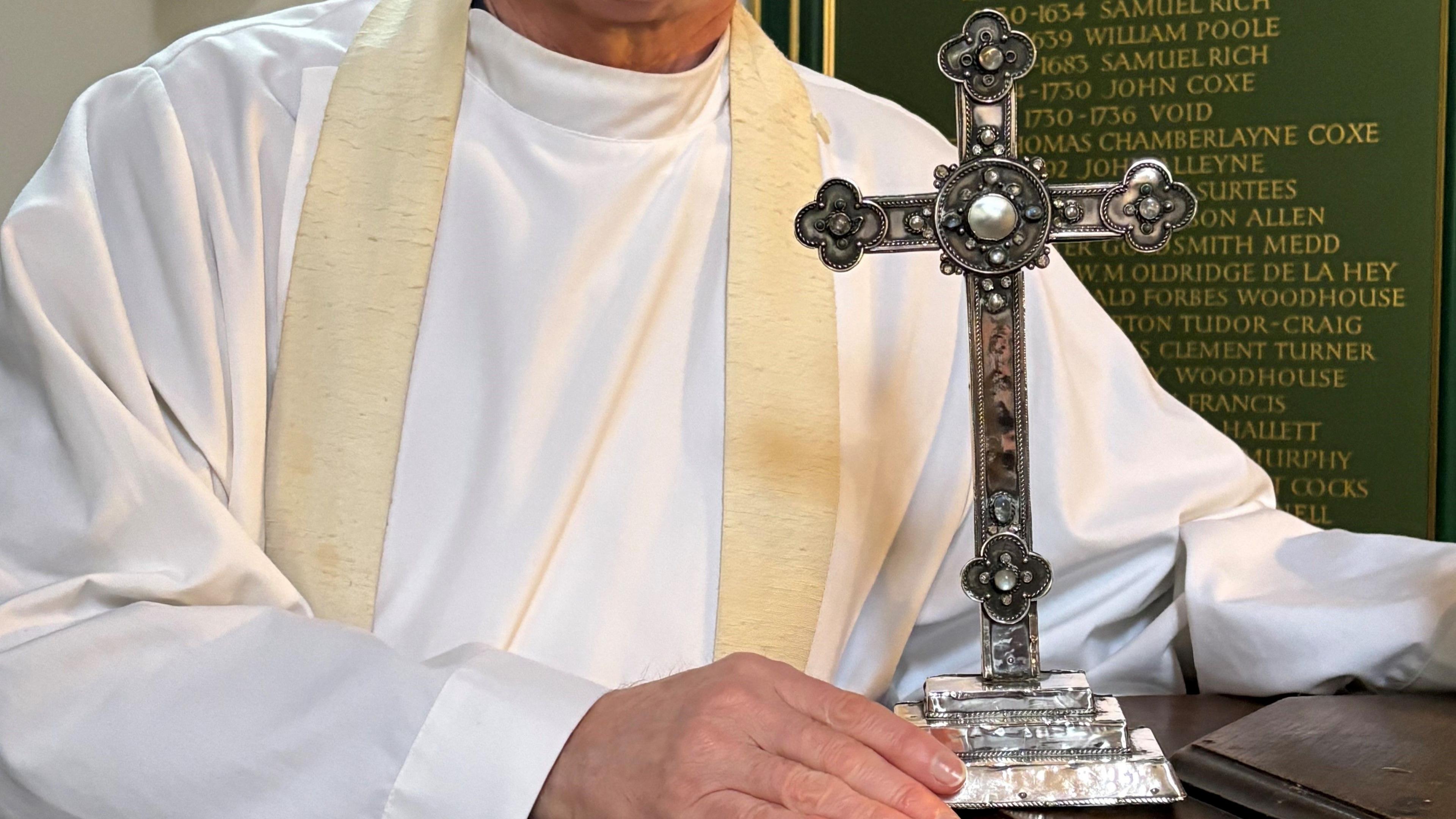 A close-up picture showing the Reverend David Minns holding the base of the silver cross, which is sitting on top of a stone altar. The cross is intricately engraved with a flowery and dotted pattern.