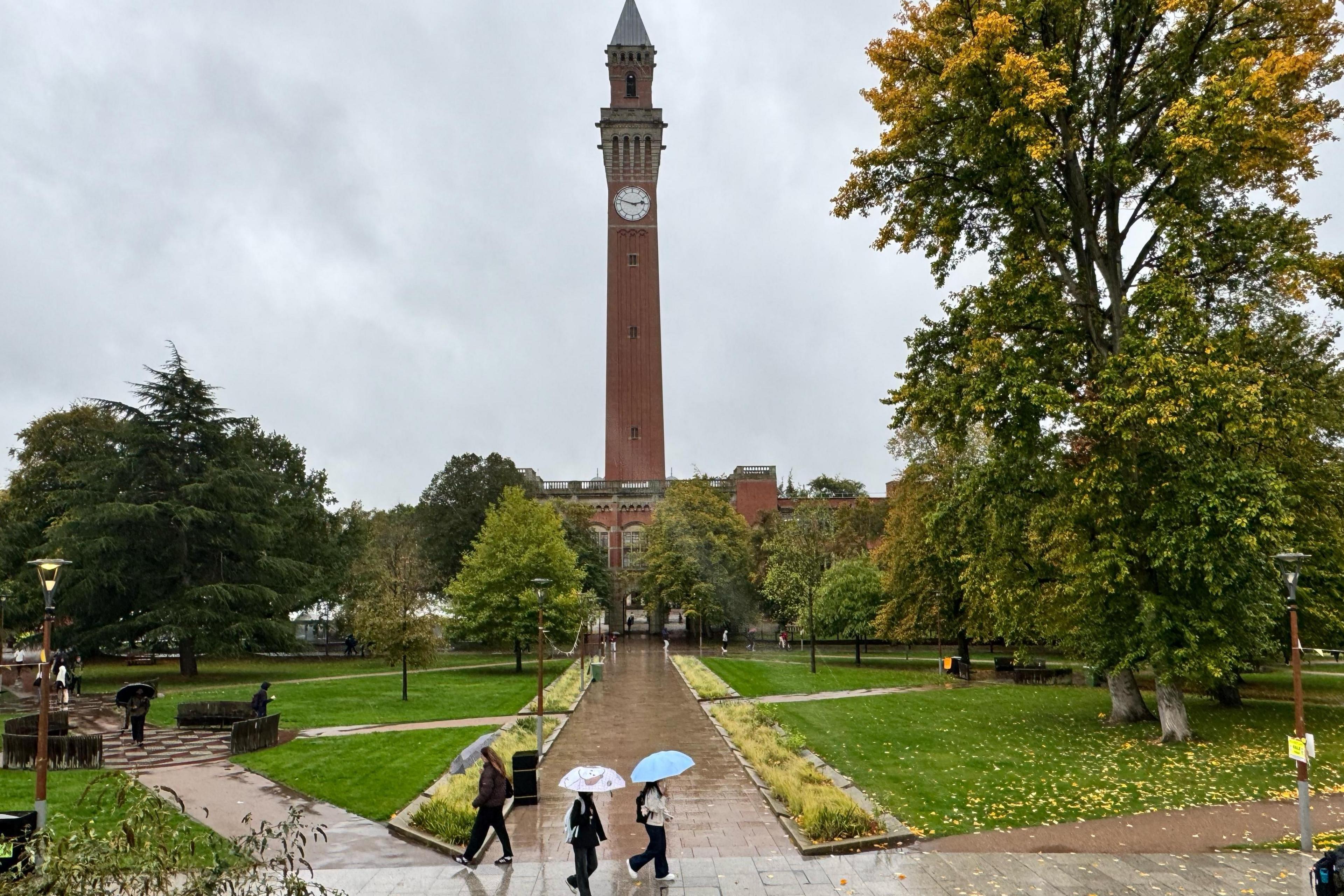 A city park with a few scattered pedestrians, many carrying umbrellas. A number of mature trees are visible, one with yellowing autumn leaves starting to dot the grass. Behind, a tall, self-standing red-brick clock tower with a white face dominates the scene.