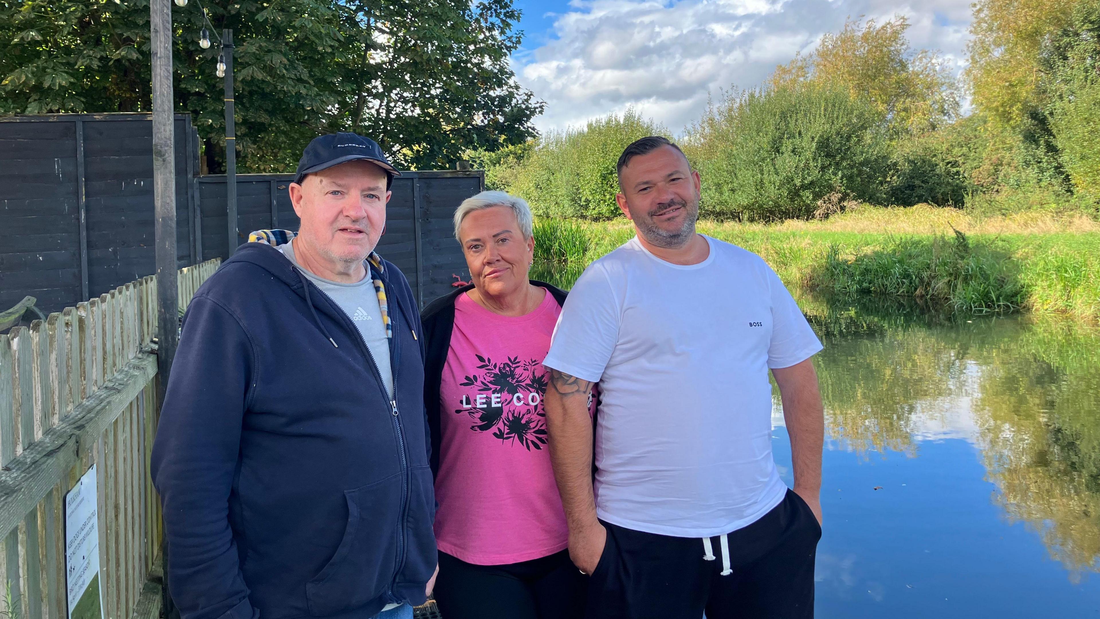 Ben Stimpson, Helen Walker and Tom Christy smiling for camera. Ben is wearing a blue zip-up hoodie and blue cap. Ms Walker is wearing a pink T-shirt and Mr Christy is in a white T-shirt. They are standing in front of a large area of water with trees and greenery in the background. 