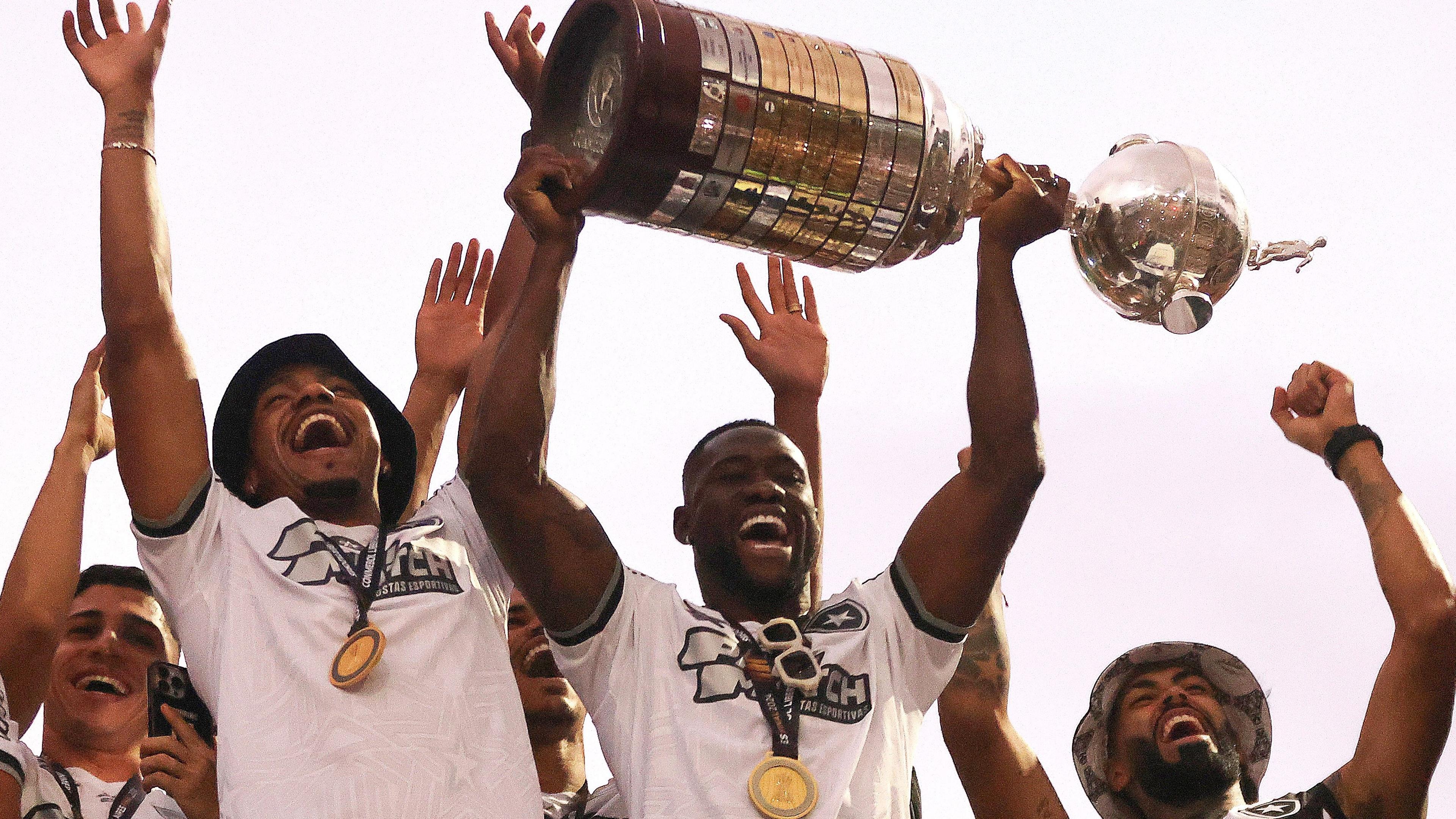 Bastos, wearing a white T-shirt with a gold medal around his neck, lifts the Copa Libertadores trophy above his head while surrounded by four other people lifting their arms in celebration