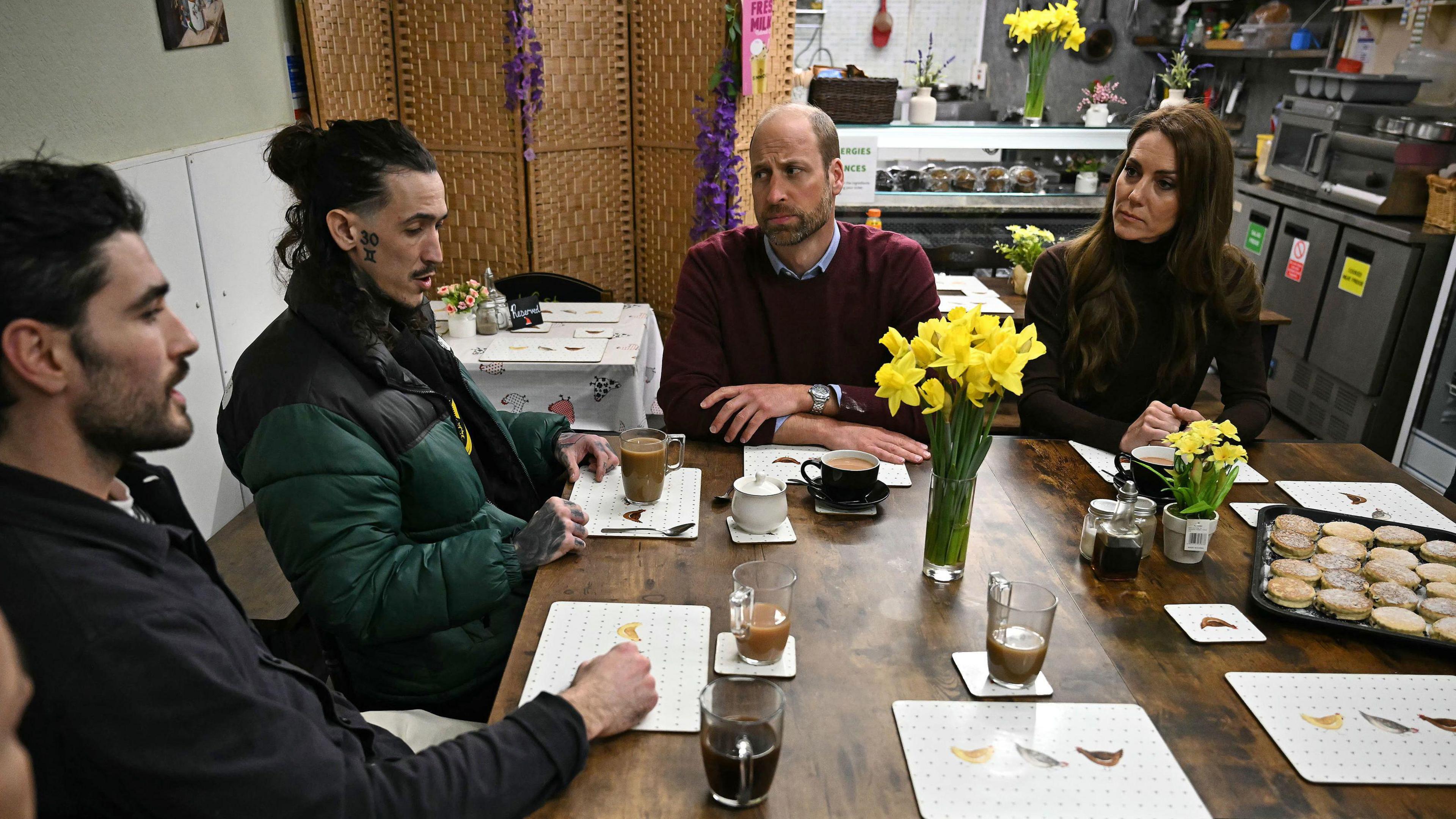 Prince and Princess of Wales sit around a wooden table with bunches of daffodil on in jugs. Two men sit on the left with cups of tea. The man closest to William is talking, he is wearing a green Puffa jacket and has his hair pulled back in a bun, while the man who sits next to him has short dark hair and is wearing a black shirt. Welsh cakes are on the table.