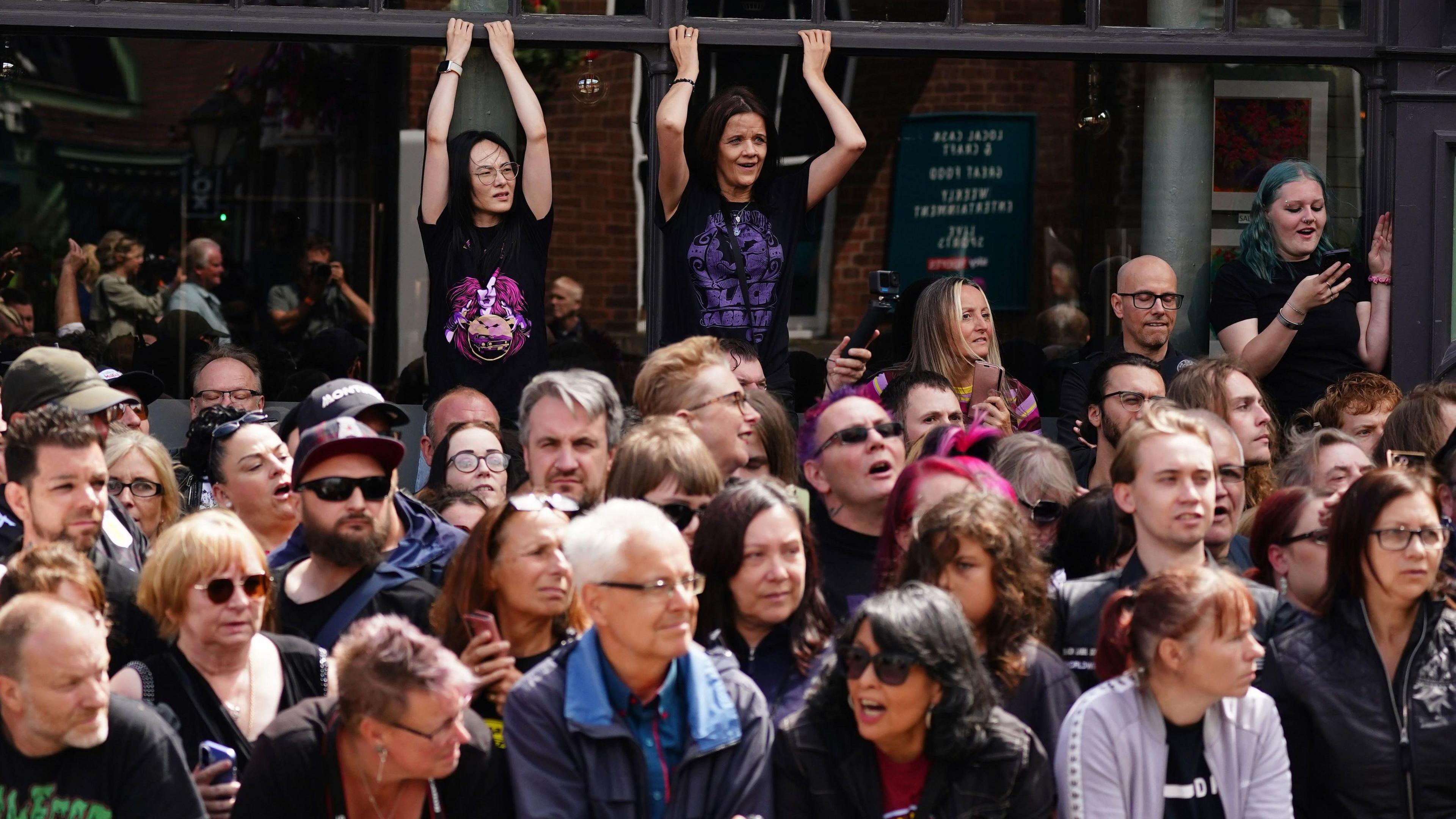 Dozens of people gather in a crowd on a pavement. Above them are two women standing on something, elevating them above everyone else. They are holding onto the outside of a building and are wearing black and purple Black Sabbath t-shirts.