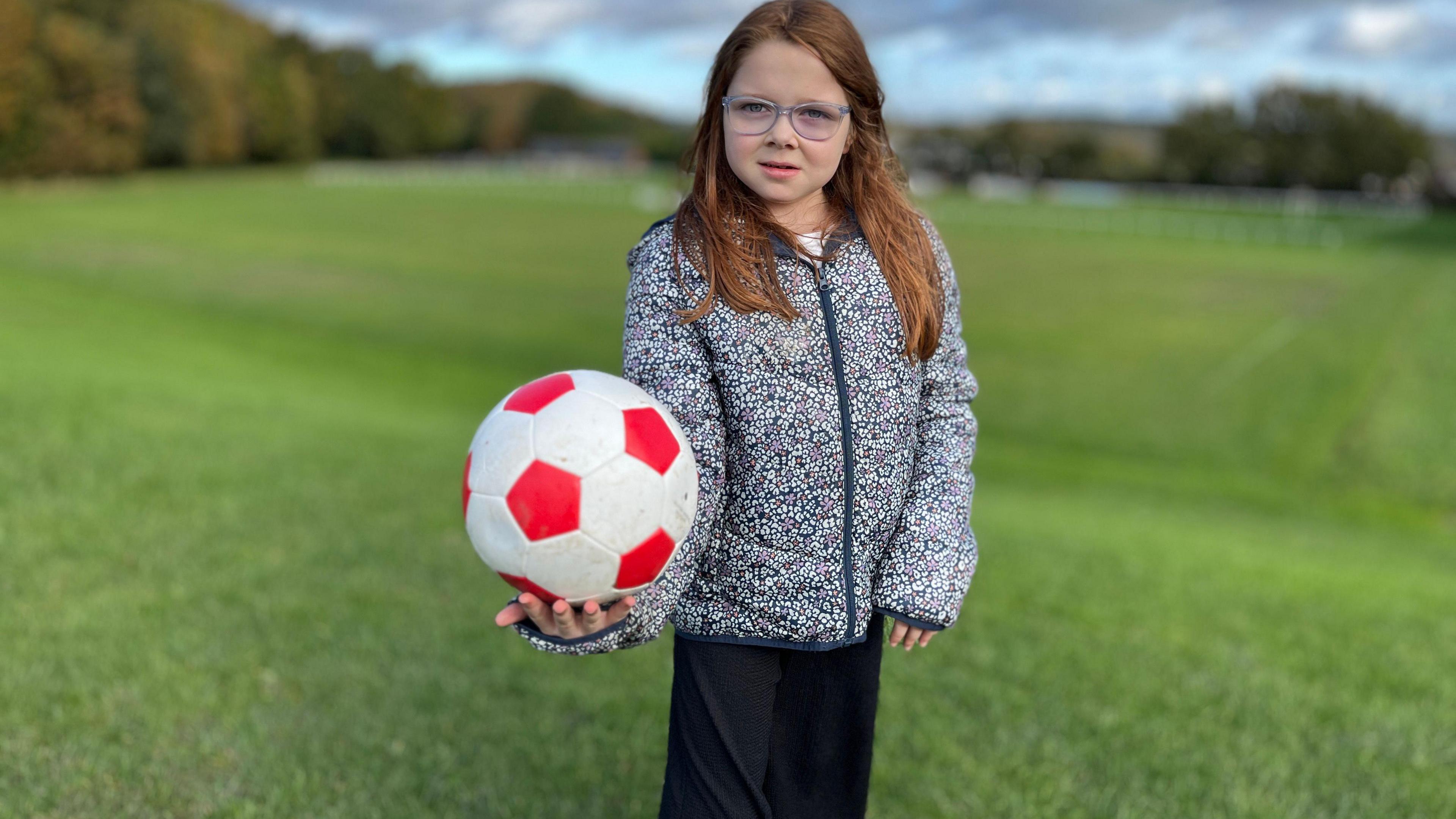 A girl wearing a grey and purple flower jacket, black trousers and glasses, holding a red and white football in one hand. Grass and trees are in the background.