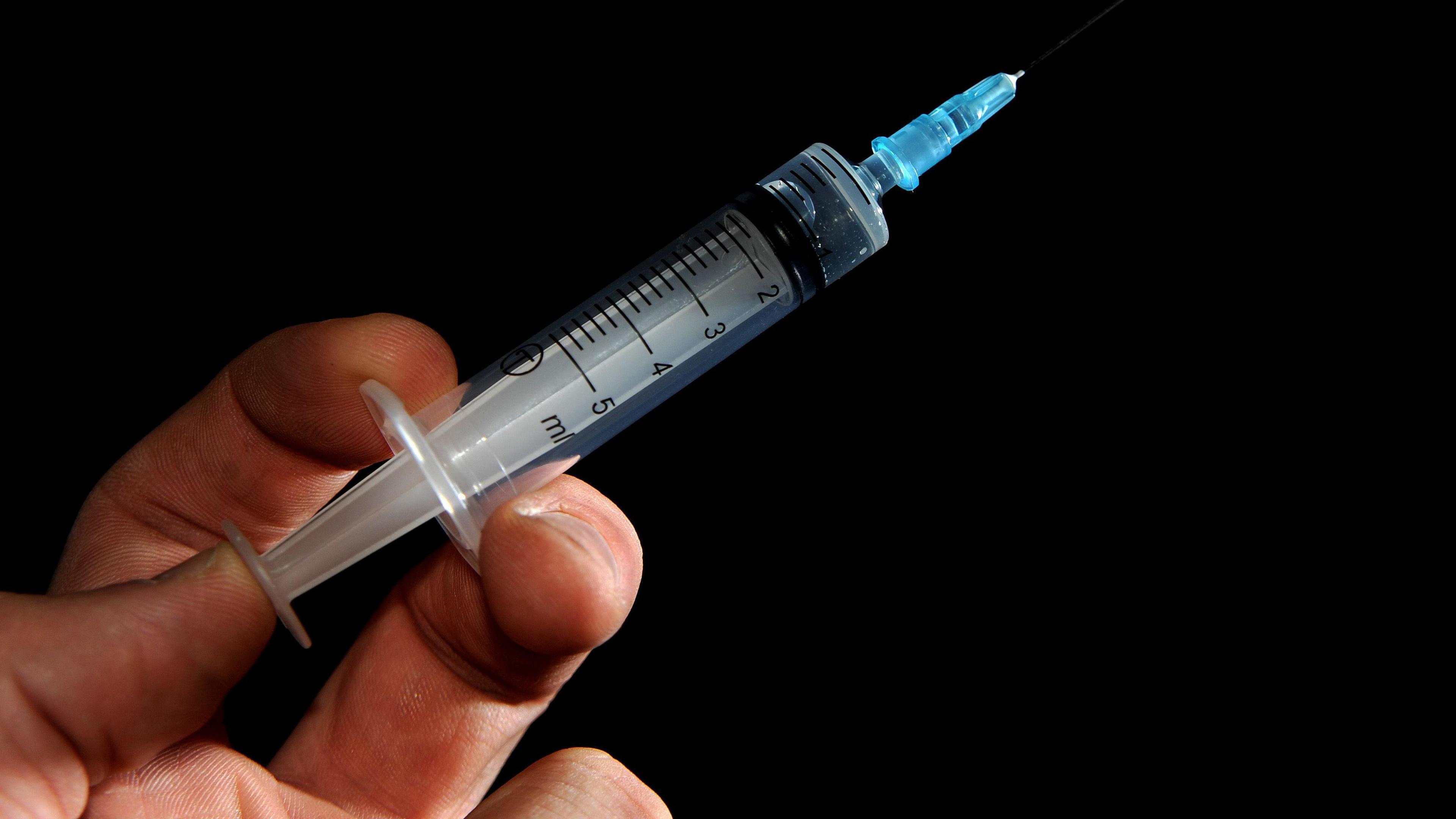 A hand holding a syringe with a small amount of clear liquid. The needle is held by a blue plastic surround, and the image is taken against a black background.