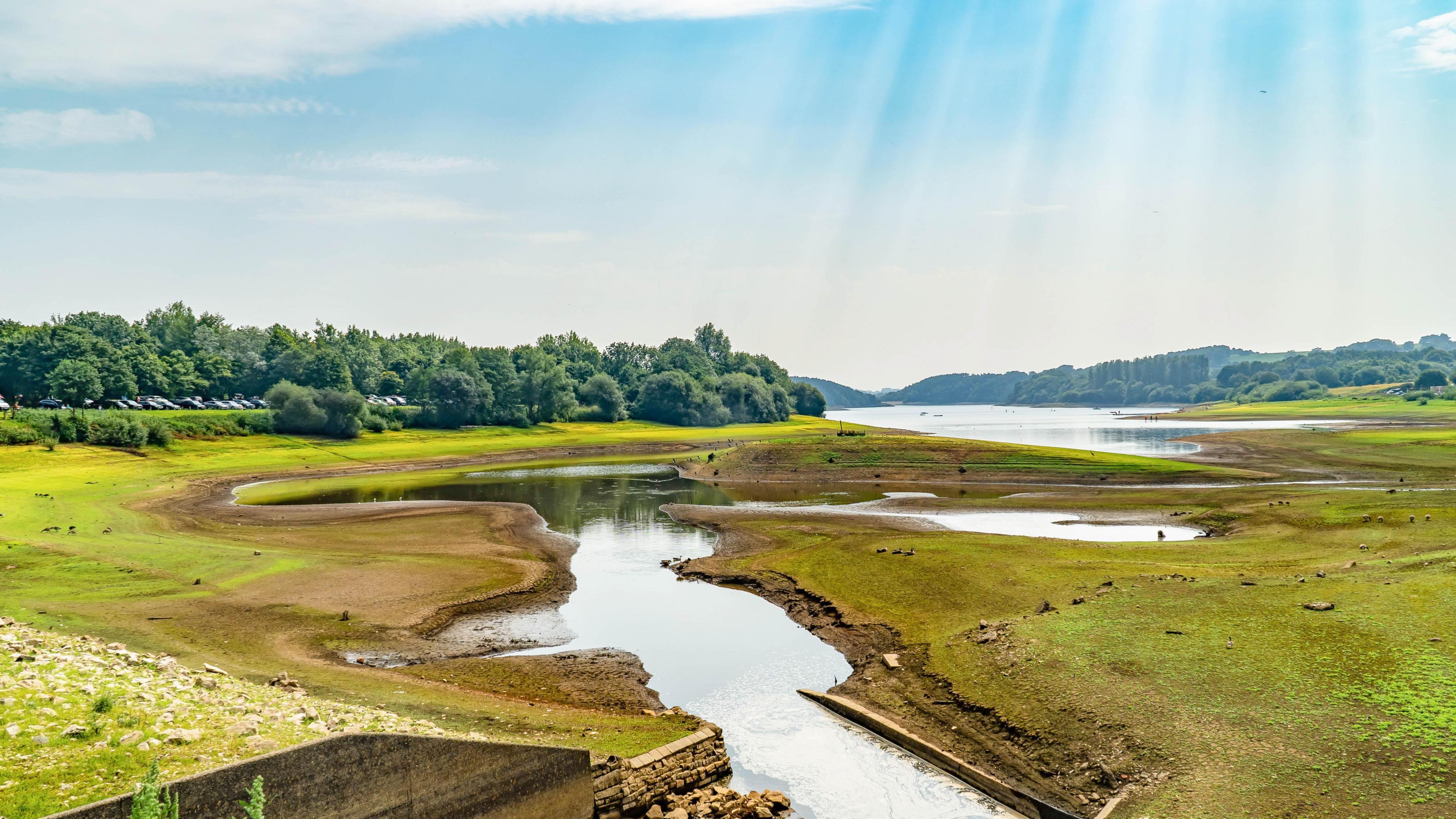 A large lake in Meerbrook, with grass covered fields dipping down to hte water on all sides. A forest is on the left hand bank and again in the extreme distance