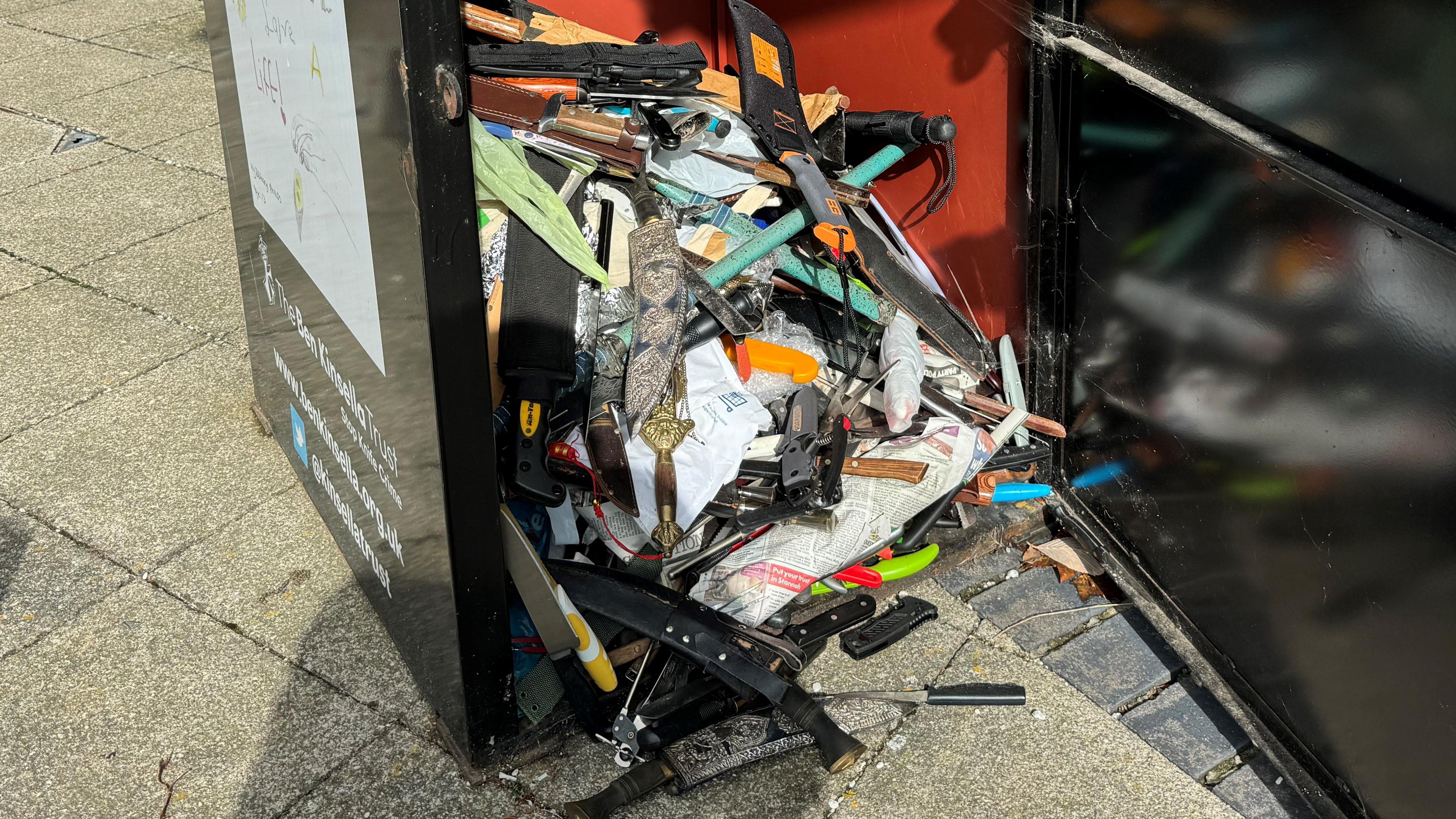 An open knife bin outside a police station with several blades spilling out onto the pavement.