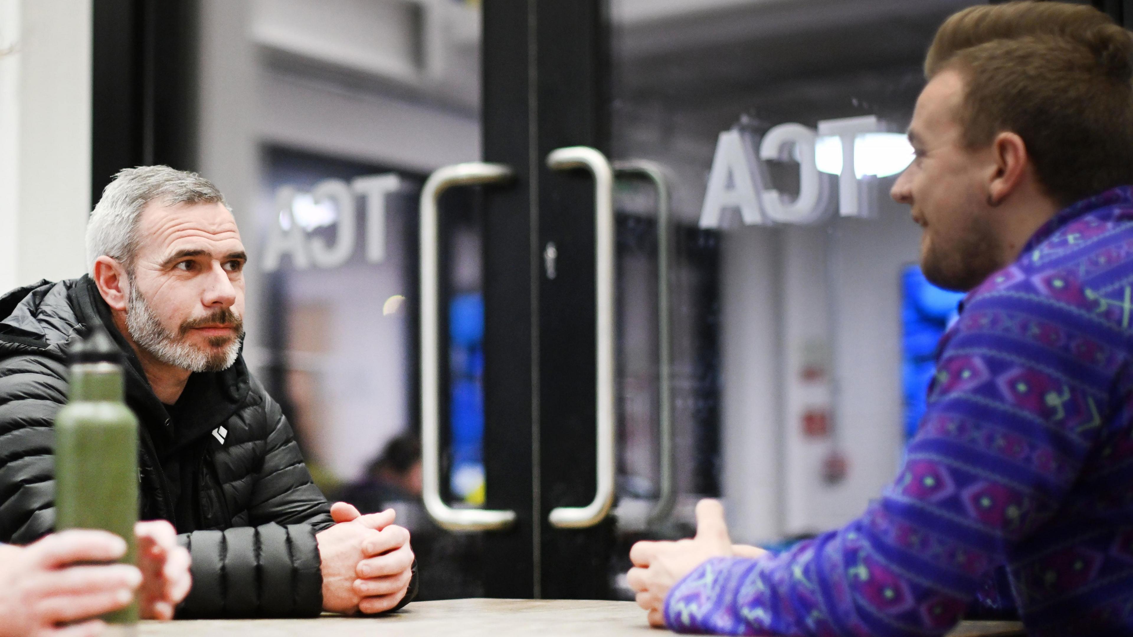 Two men are sat opposite eachother at a table. The man on the left is in focus, he has grey hair and a grey beard and is wearing a black coat. The man on the right is out of focus, only the side of his face is on show. He has brown hair and is wearing a purple patterned jumper. Both men have their hands on the table. Behind them is a large glass door