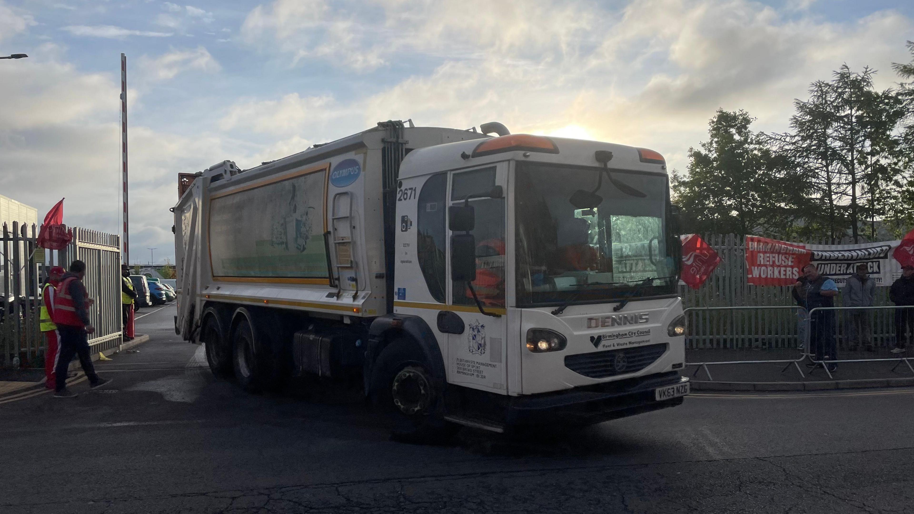 A large white bin lorry driving out of the exit of a depot. There are large metal fences surrounding it, with red unite the union flags on them.