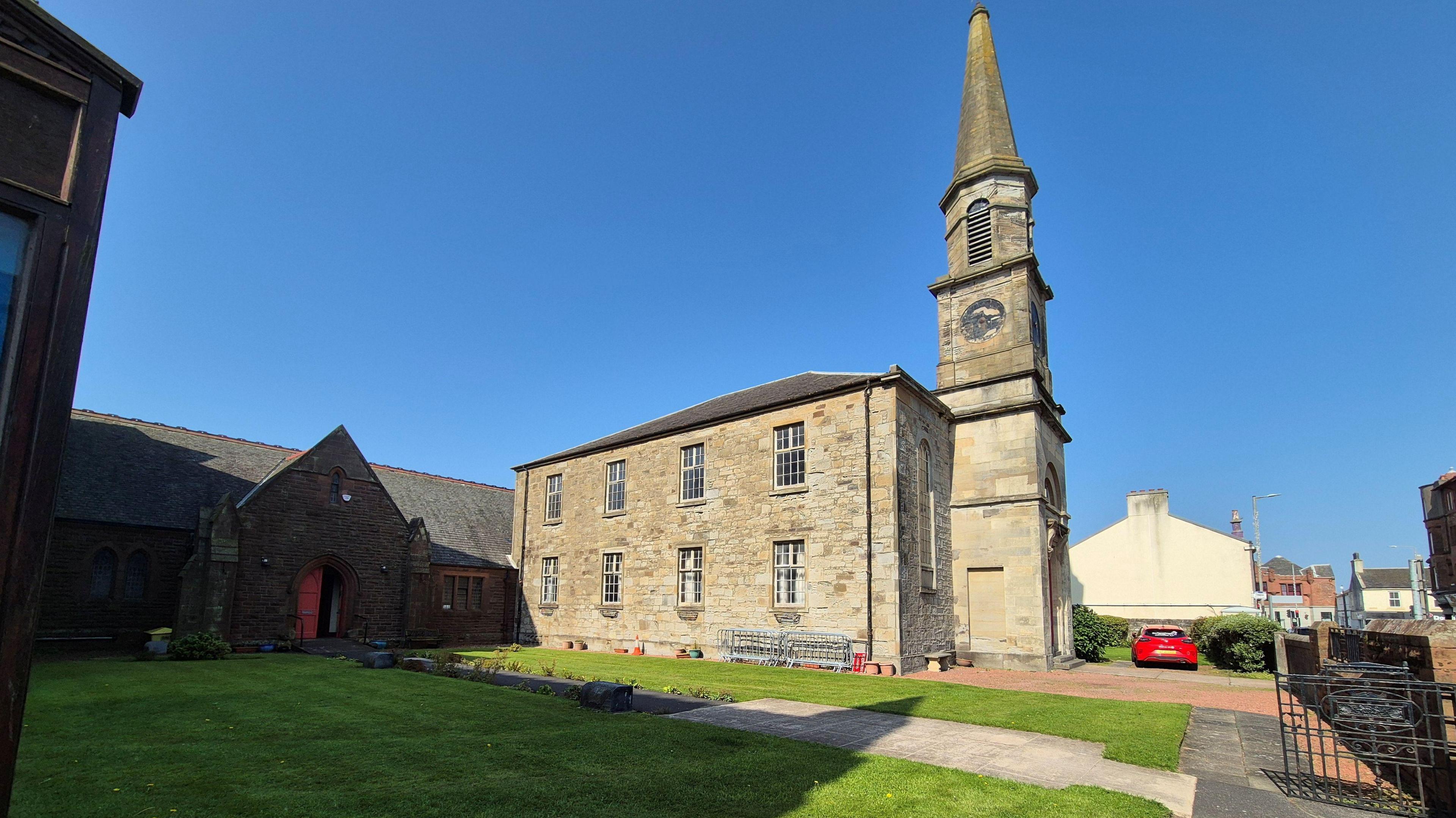 An outdoor view of part of a church building. It is a sandstone build bathed in sunlight and has a tall steeple.
