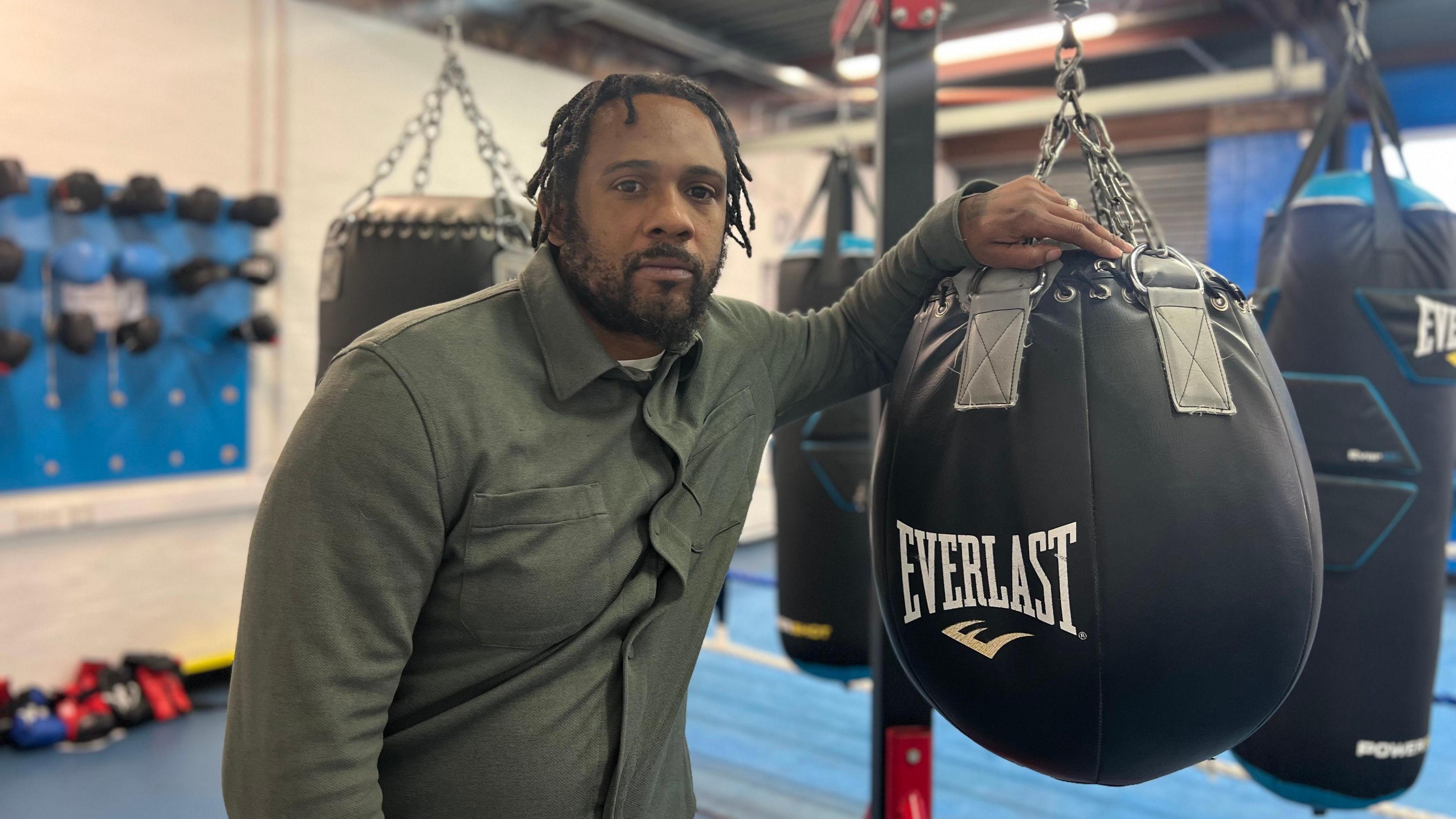 Dayton is standing next to a punching bag in a boxing gym. He has a green shirt on and wears his hair in braids. 