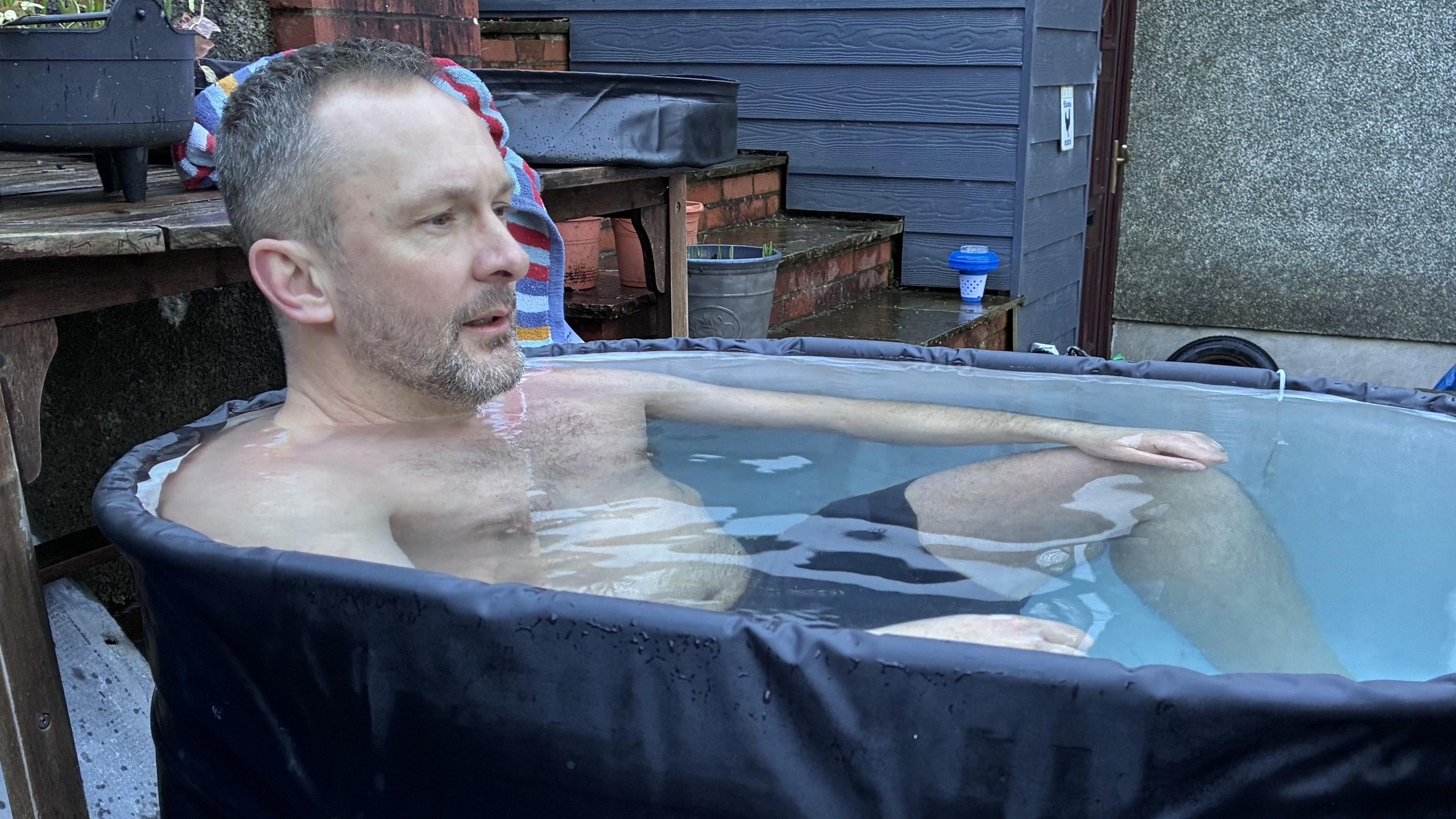 Gerwyn yn eistedd mewn bath ia mewn siorts nofio, yn edrych yn anghyfforddus