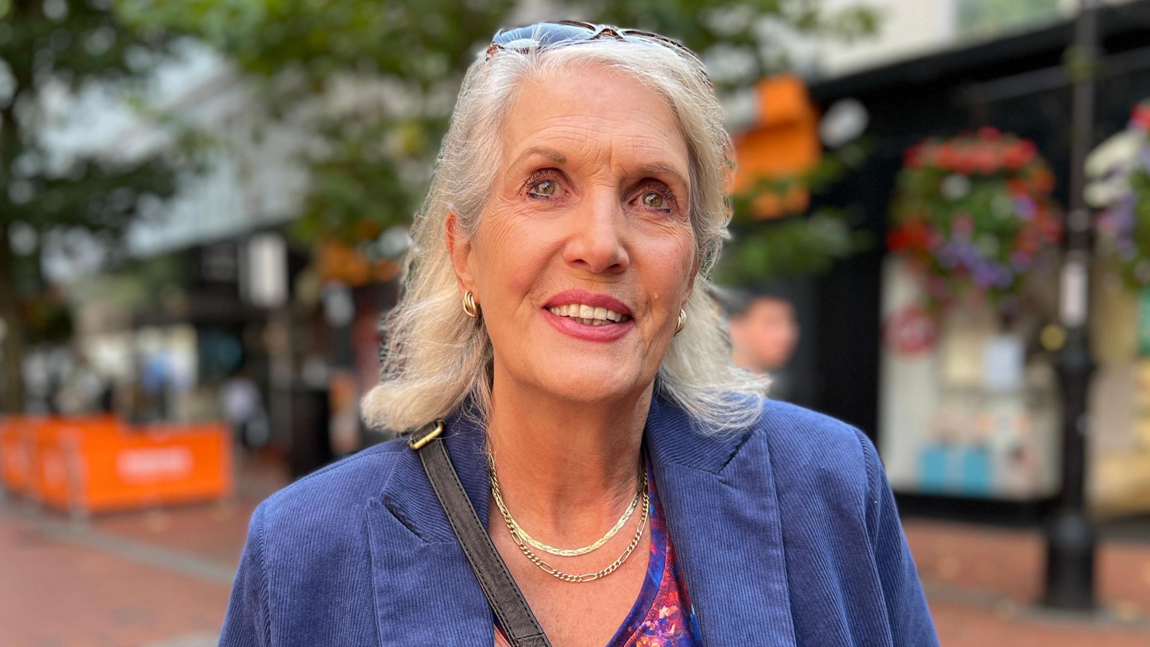 Gwendoline Hurn is smiling and wearing a blue blazer jacket with a black bag strap over her shoulder. She has light blonde hair and sunglasses balanced on her head.