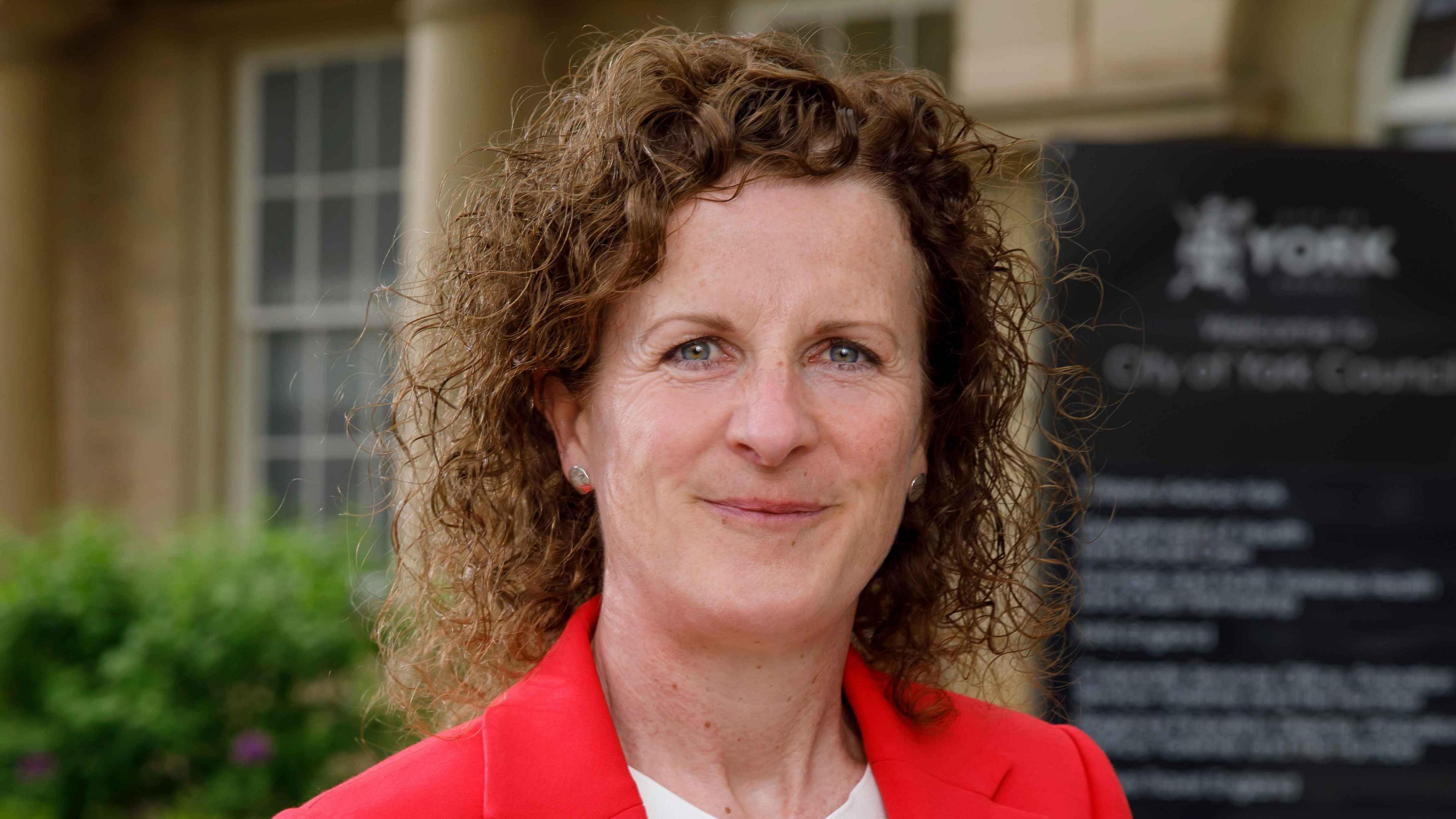 York Council leader Claire Douglas: a woman with curly dark hair and blue eyes. She is smiling into the camera and wearing a bright red jacket.