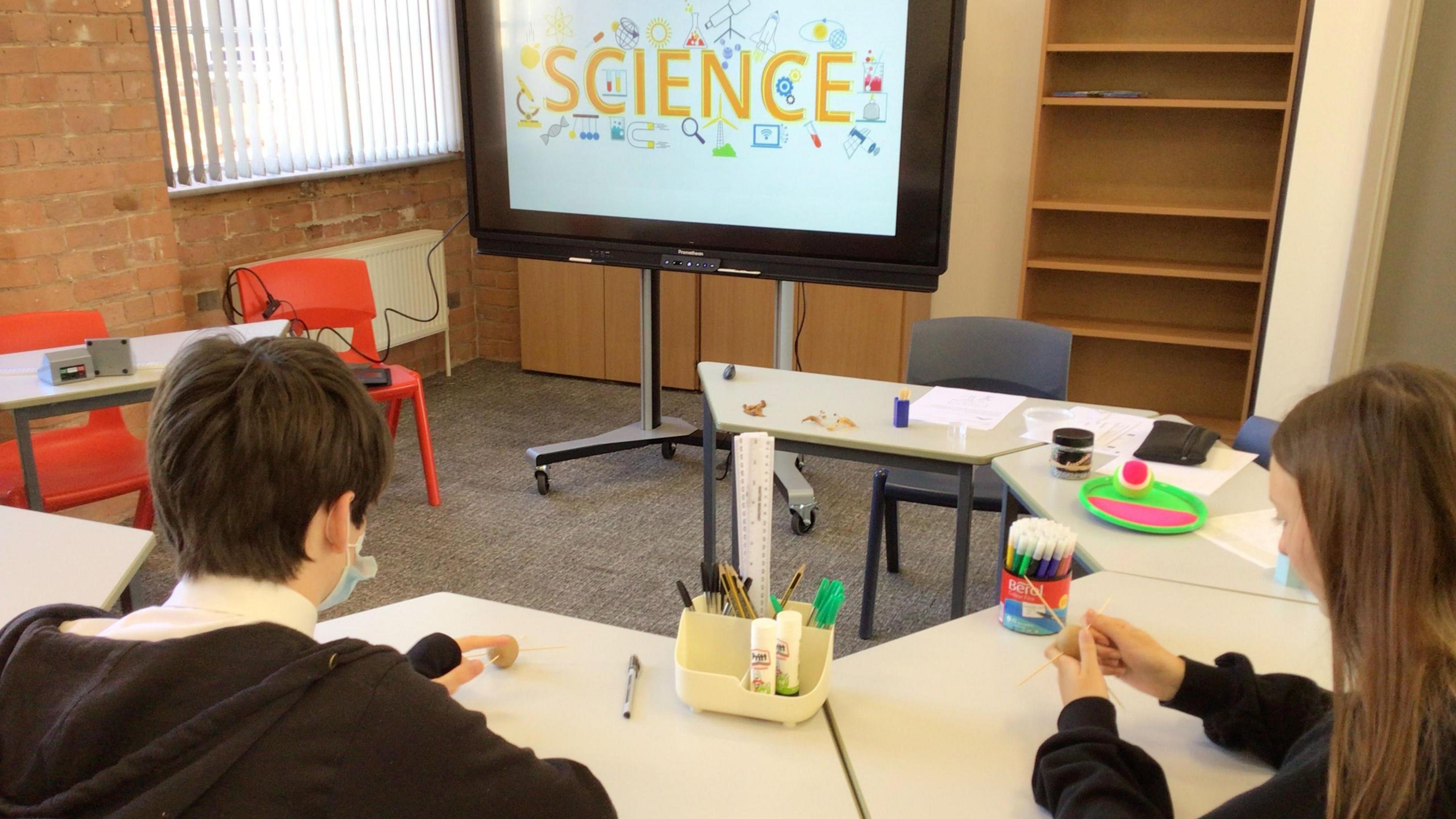 A boy and a girl sat at a ring of desks with a monitor in the background displaying the word 'science'