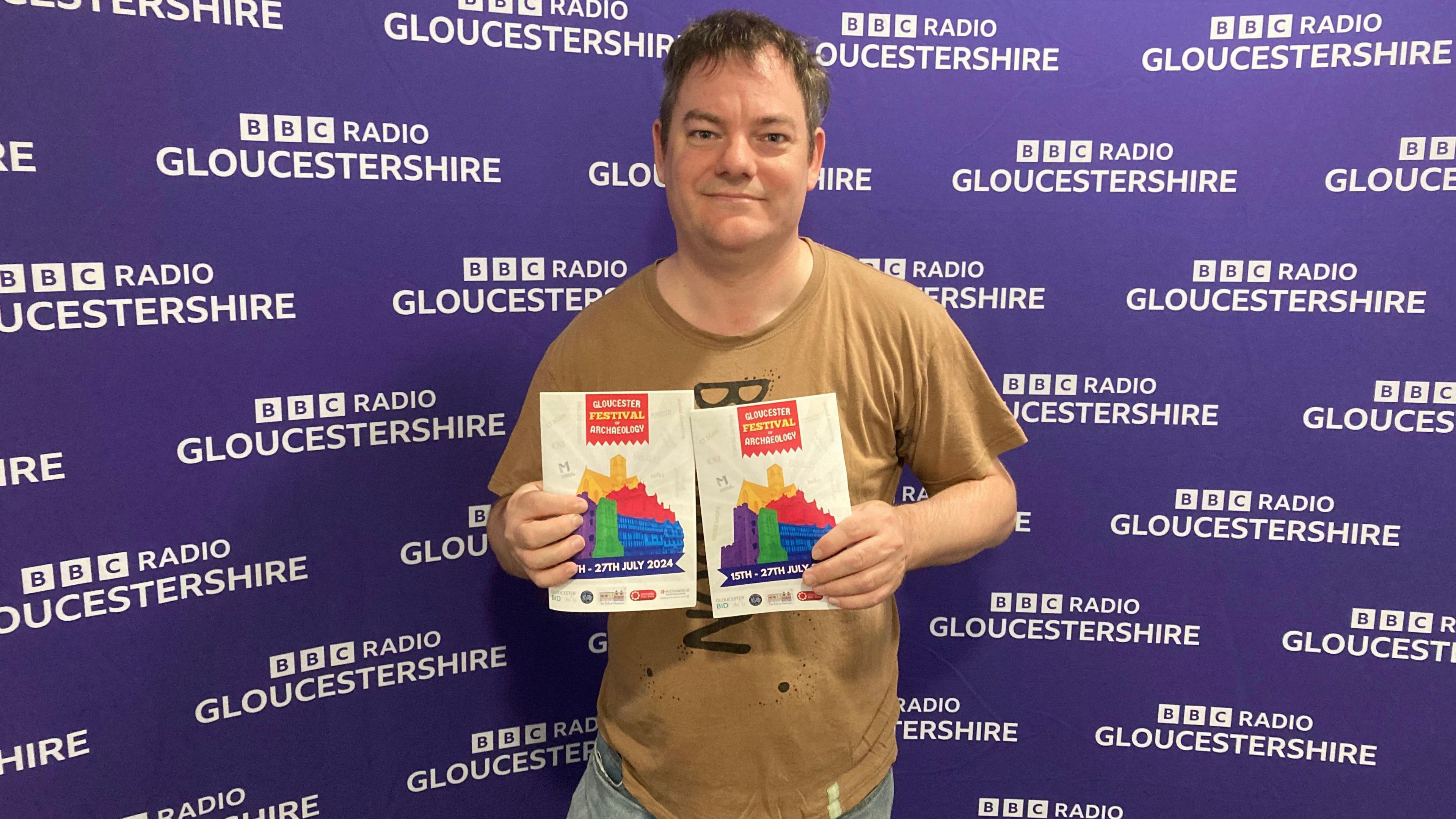 Marc Waters holding two leaflets to the festival at the BBC Radio Gloucestershire studios