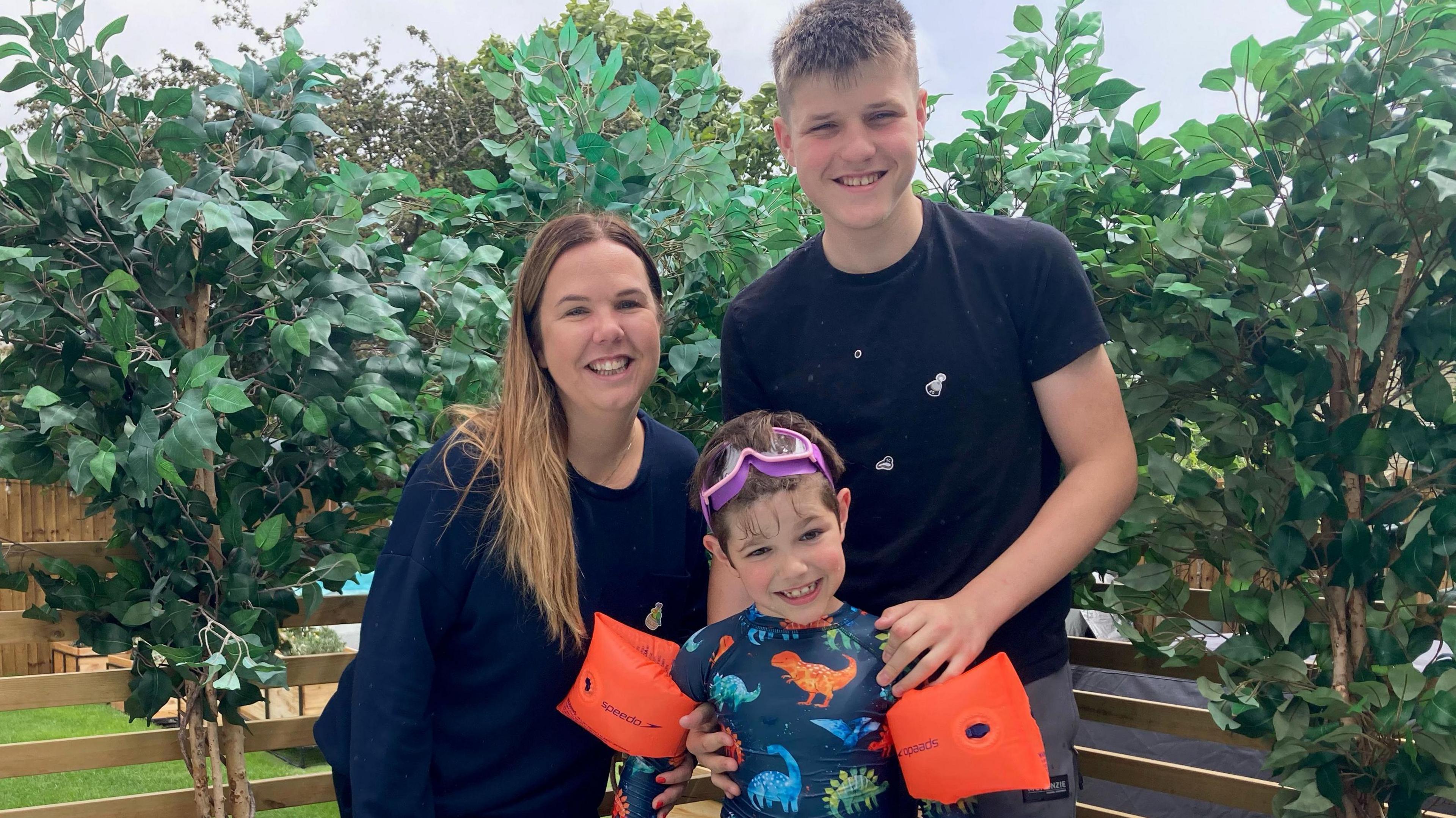 A woman with long brown hair is wearing a dark blue top and stands next to a teenage boy with short fair hair wearing a black top. Between them is a younger boy wearing purple goggles on his head a swimming top with dinosaurs on it and red water wings