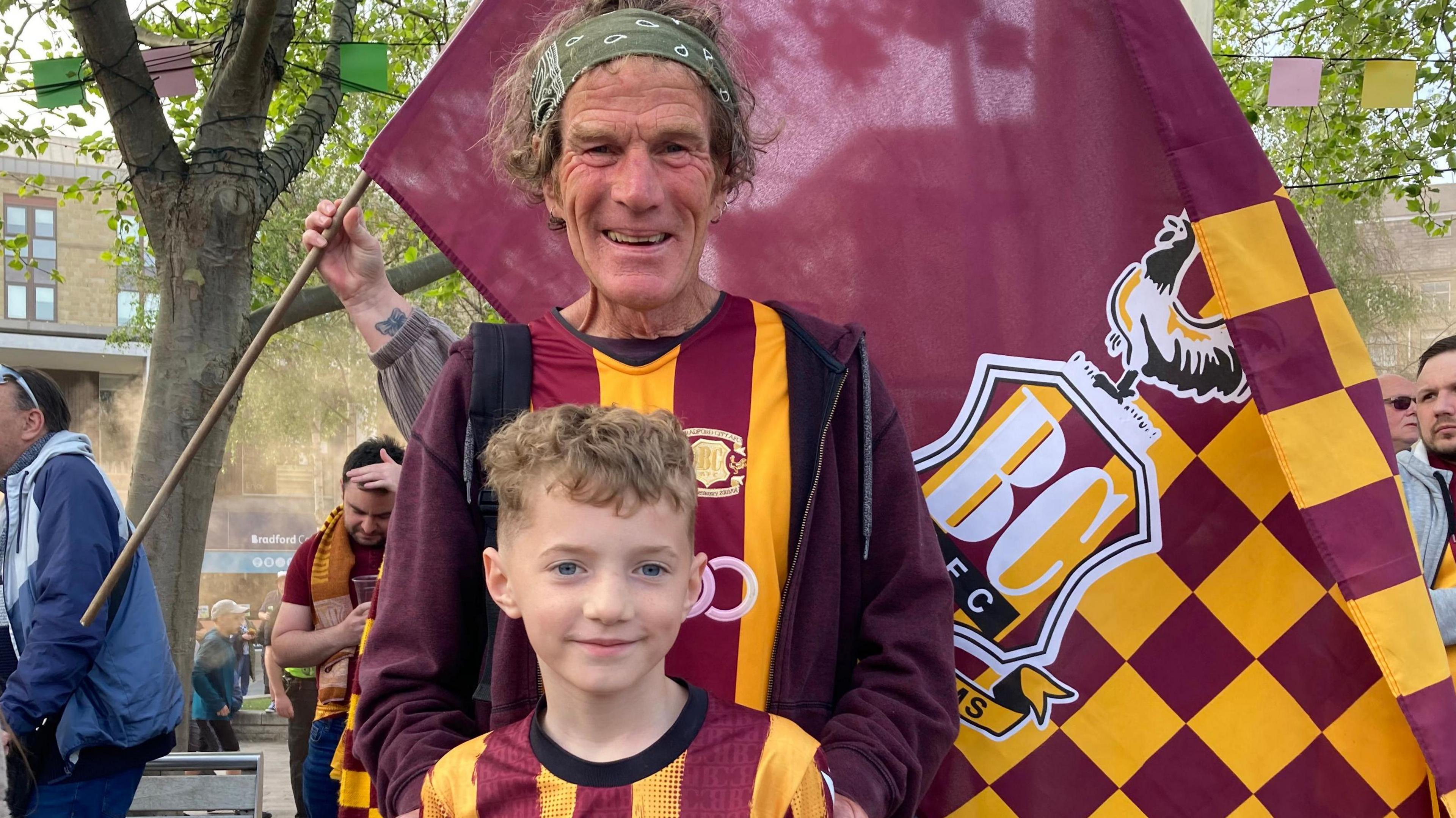 A man in his 60s with hands on boys shoulders wearing Bradford City tops smiling with flag behind them