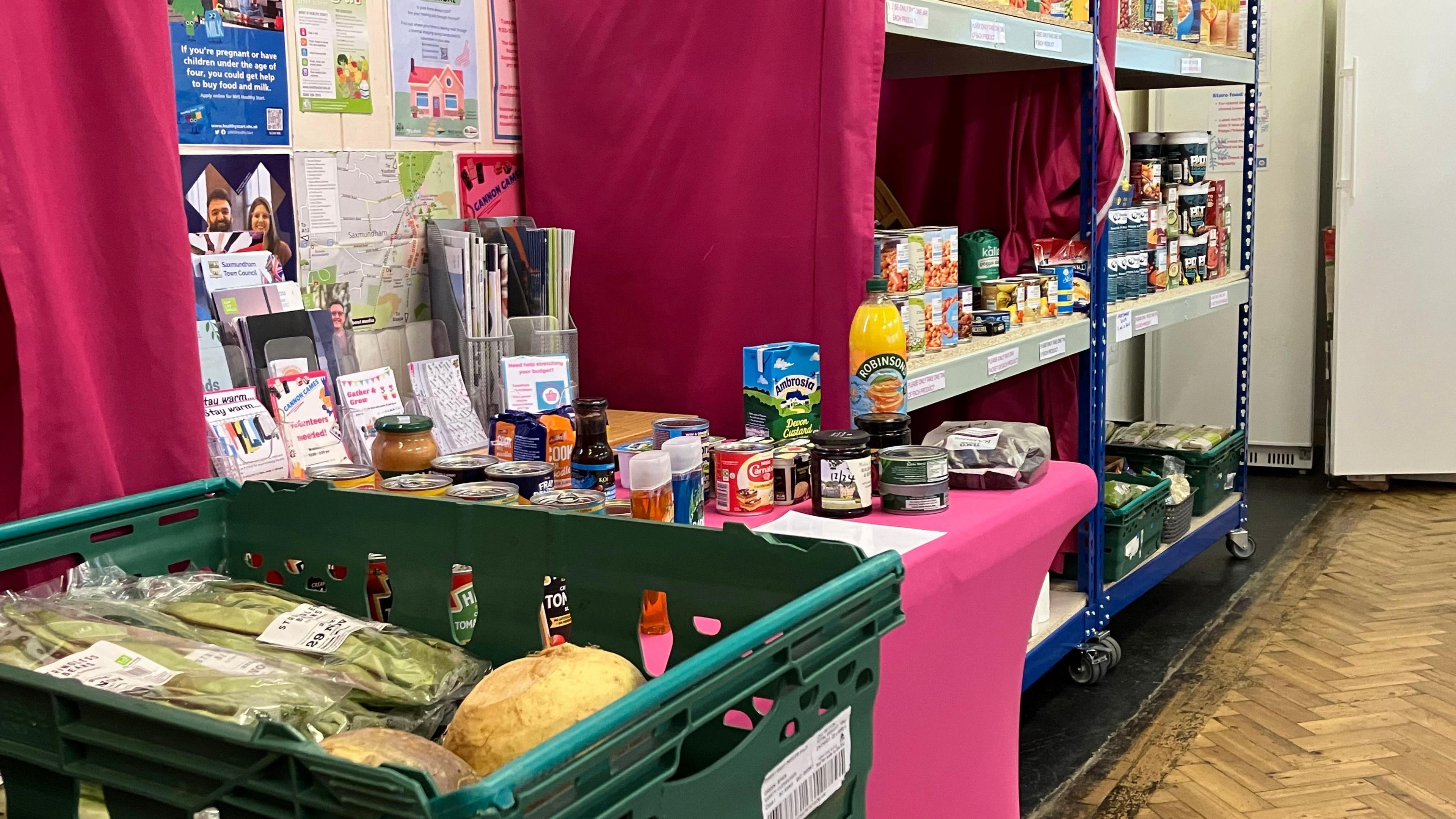 A selection of vegetables, drinks and canned foods can be seen stacked on shelves, tables and piled up in a green plastic basket