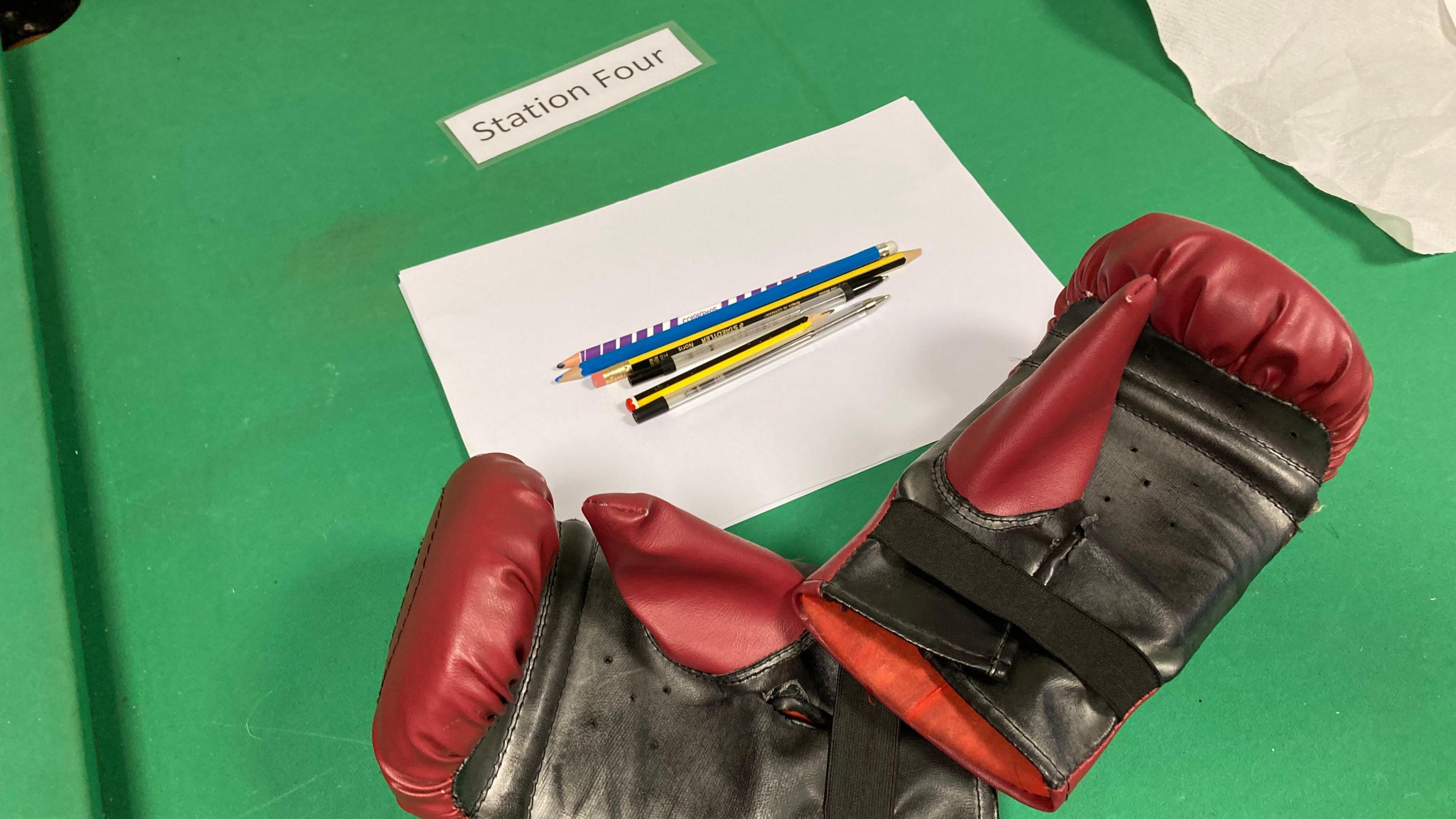 Red and black boxing gloves rest on a table. Above it a piece of paper with pencils and pens rest. 