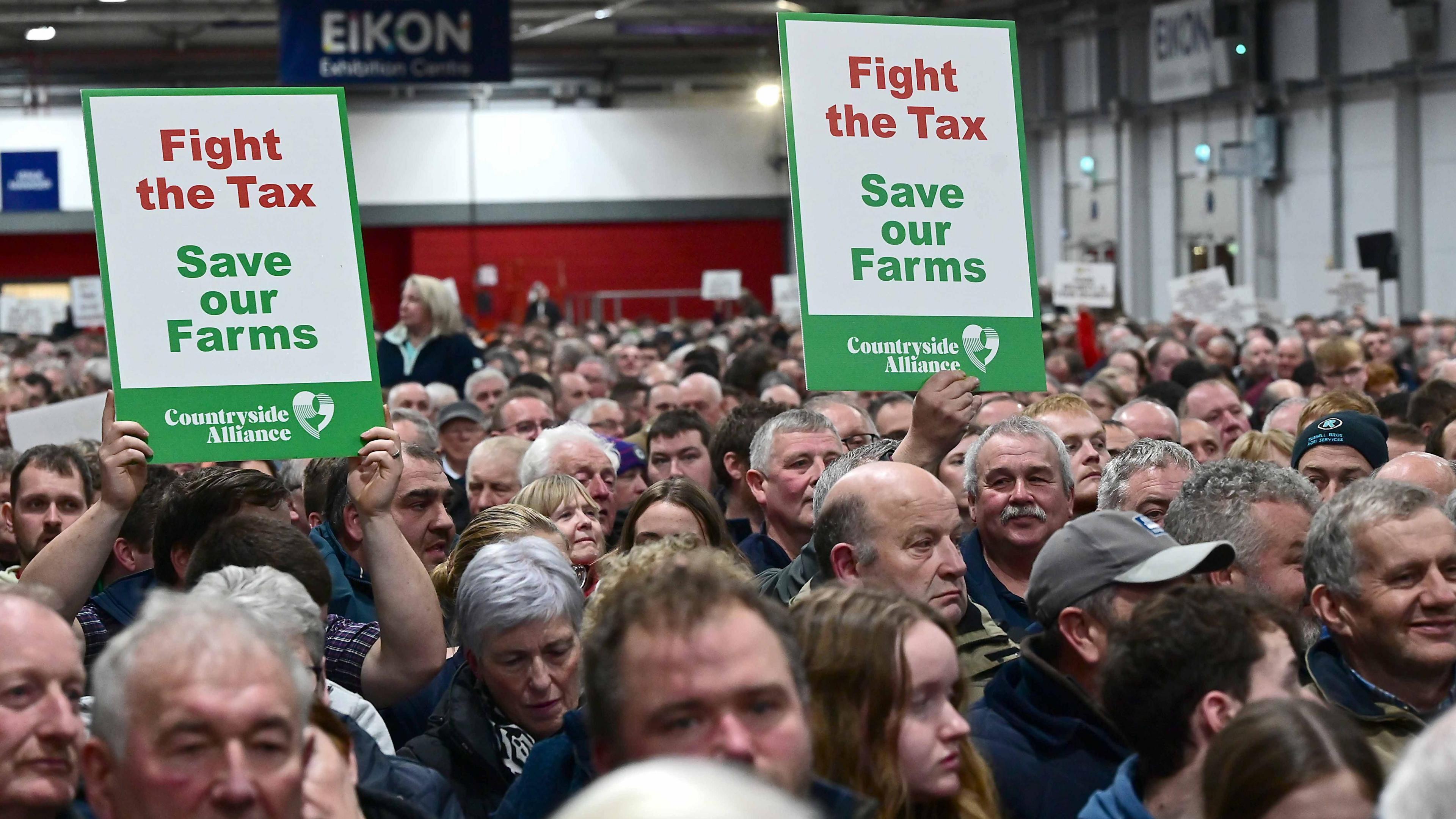 A large group of people standing close to each other. Some are holding banners that say, fight the tax, Save our farms. They are standing in a hall.