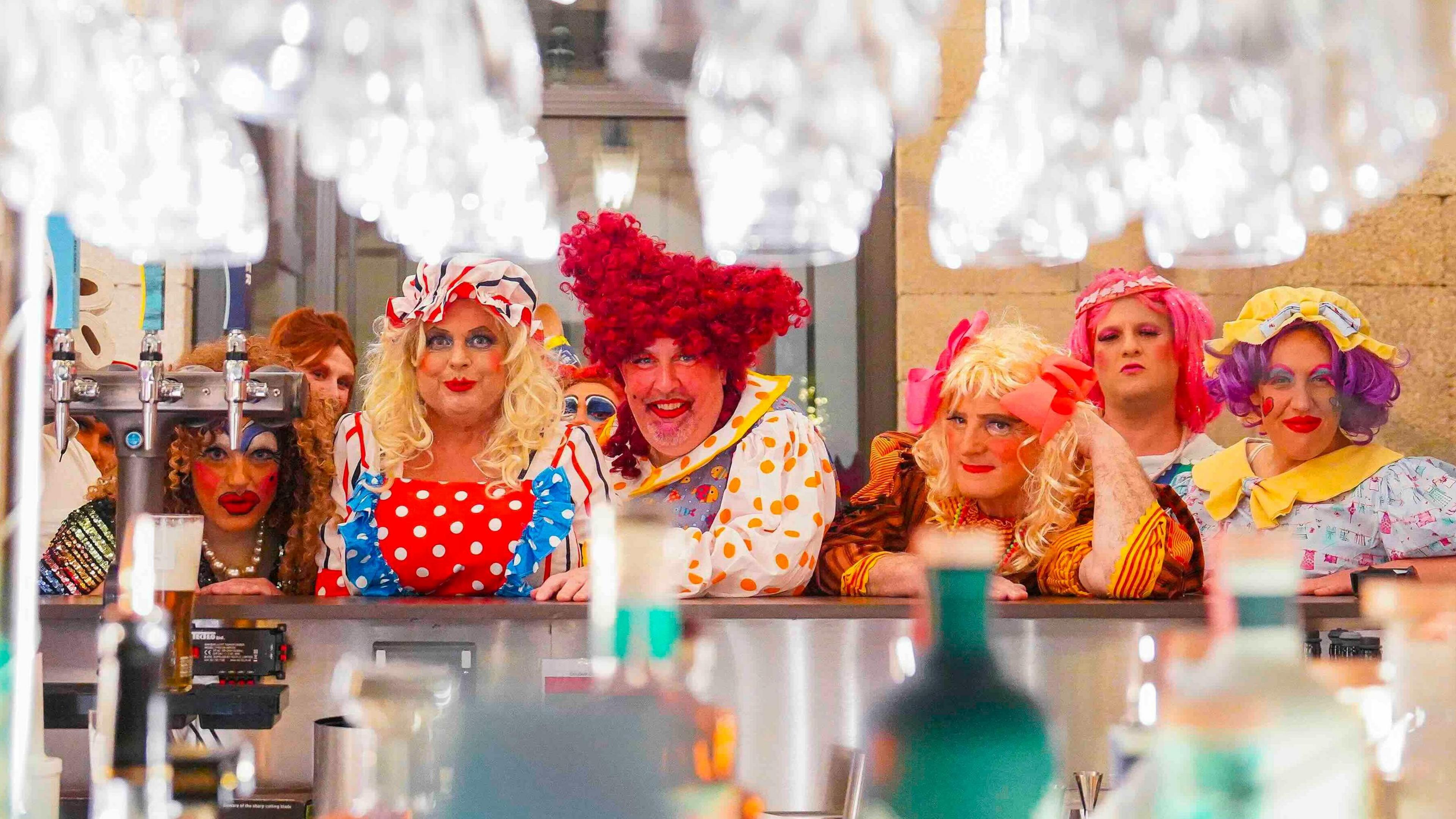 Six panto damges in bright costumes behind a bar with glasses above.