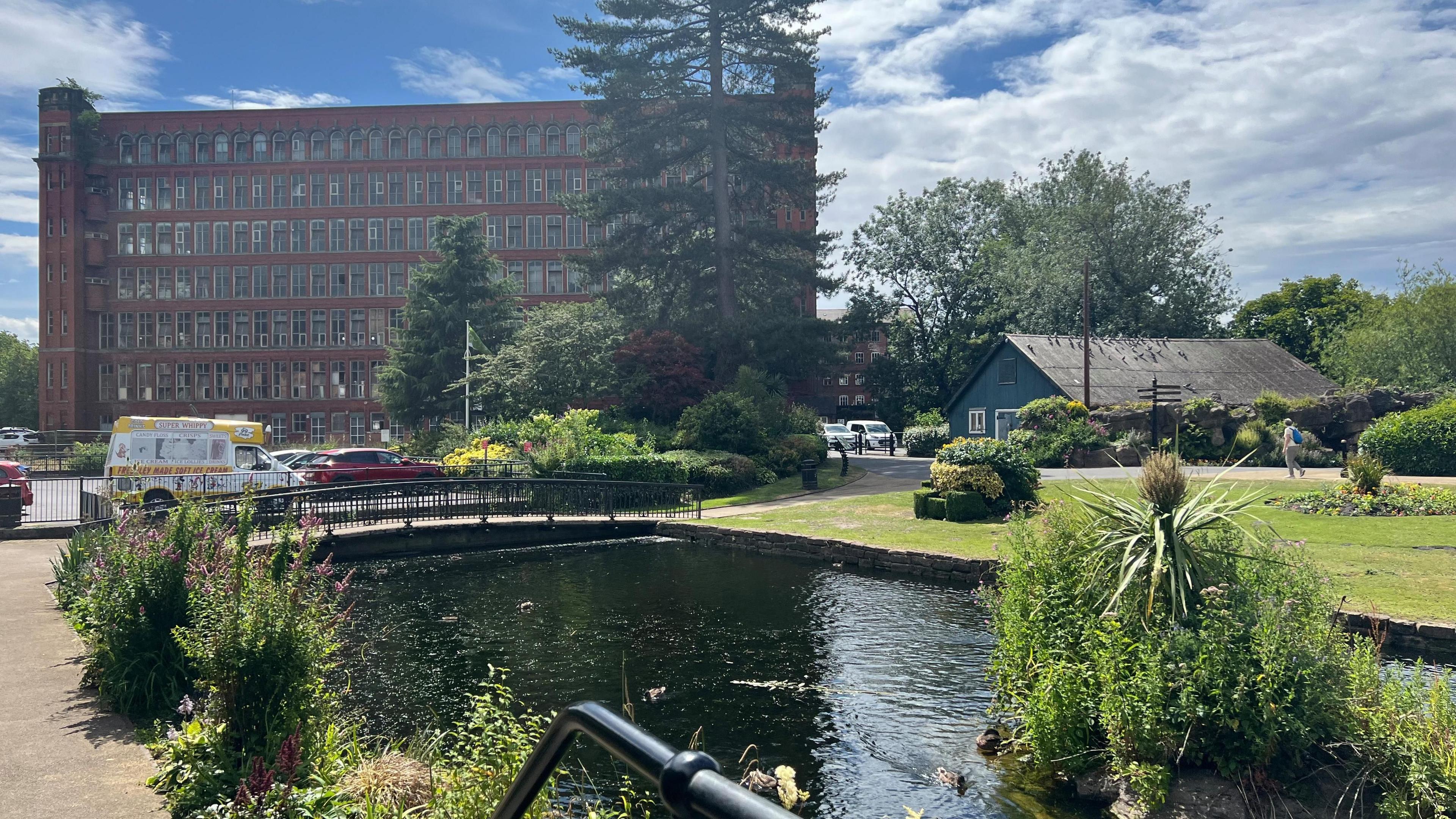 Large victorian orange brick building with numerous rectangular white paned windows infront of river with a bridge going over the top