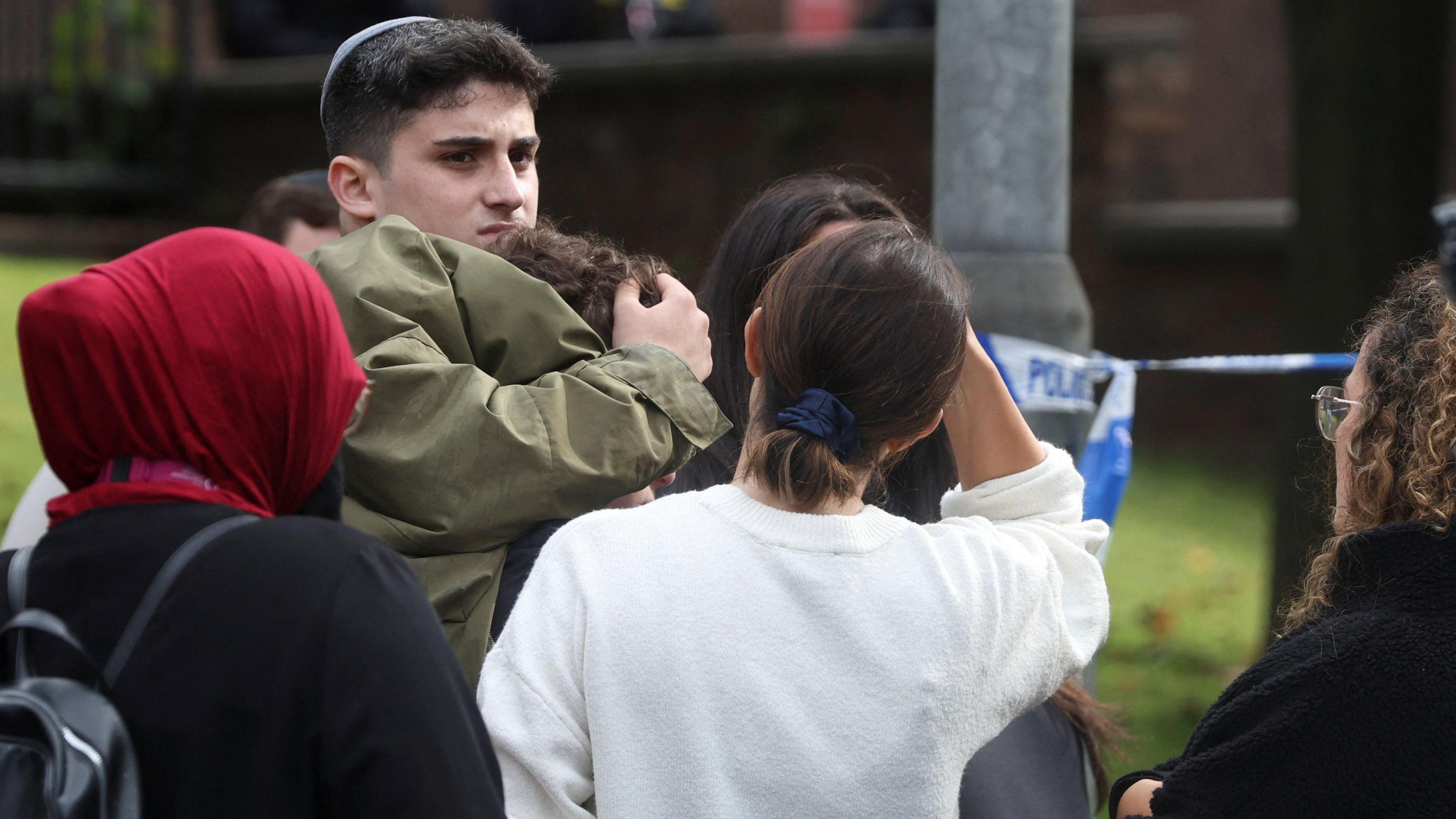 A man stands with his arm round another person, in a crowd of people standing next to police tape. A lot of their faces cannot be seen