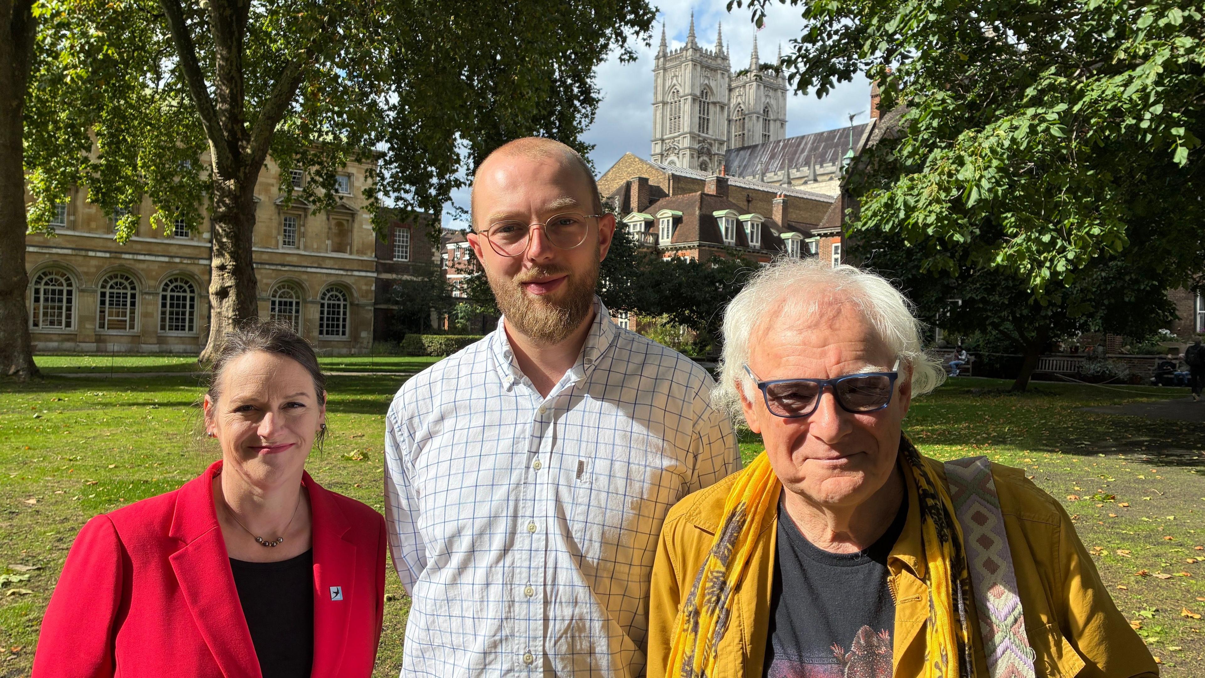 Three people smile into camera, on the left a woman in a red jacket, in the centre a taller man with a beard and glasses, wearing a white checked shirt, and on the right, a man with glasses and white hair, wearing a yellow jacket. The three of them are standing in gardens with the towers of Westminster Abbey in the background