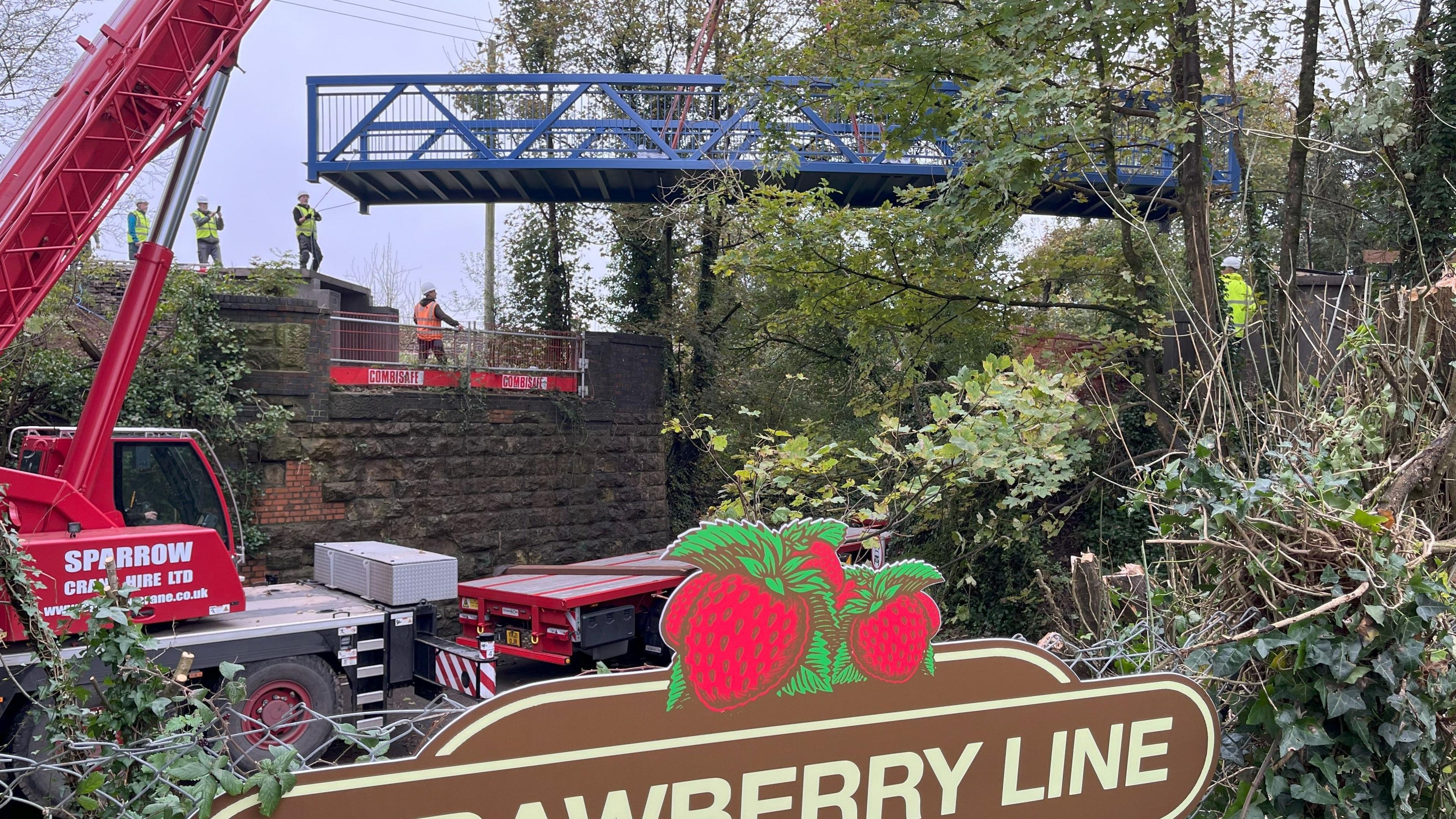 The new footbridge being lowered into place over the B3136 West Shepton In Shepton Mallet. It is a long metal bridge painted blue. Beside it is a red crane lowering it into place, with several workers in yellow hi-vis vests directing its placement. In the foreground there is a brown sign, which reads Strawberry Line with strawberries in a clump.