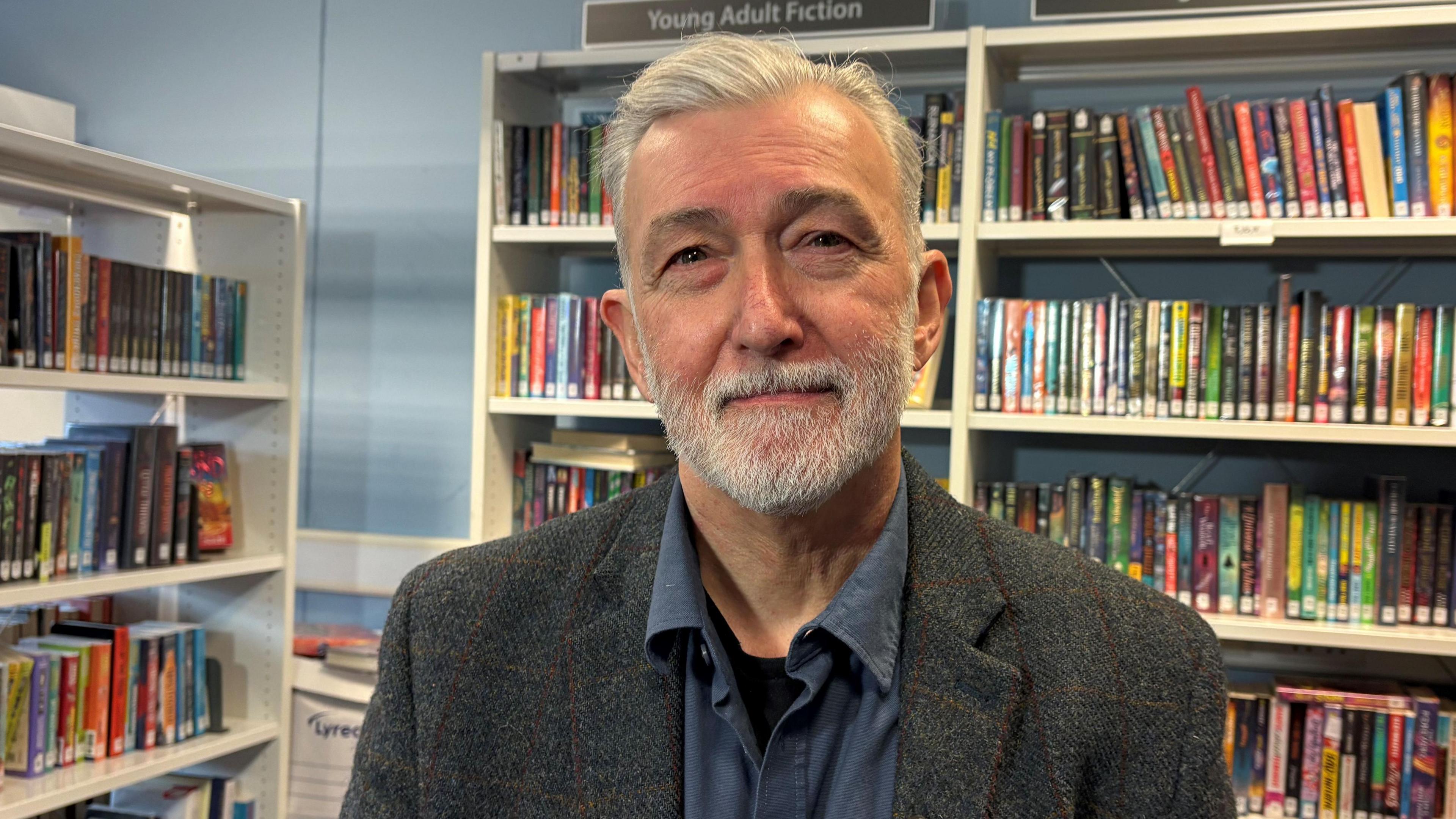 Rod McCready looking directly at the camera in a school library. He is wearign a dark grey jacket and a dark blue shirt.