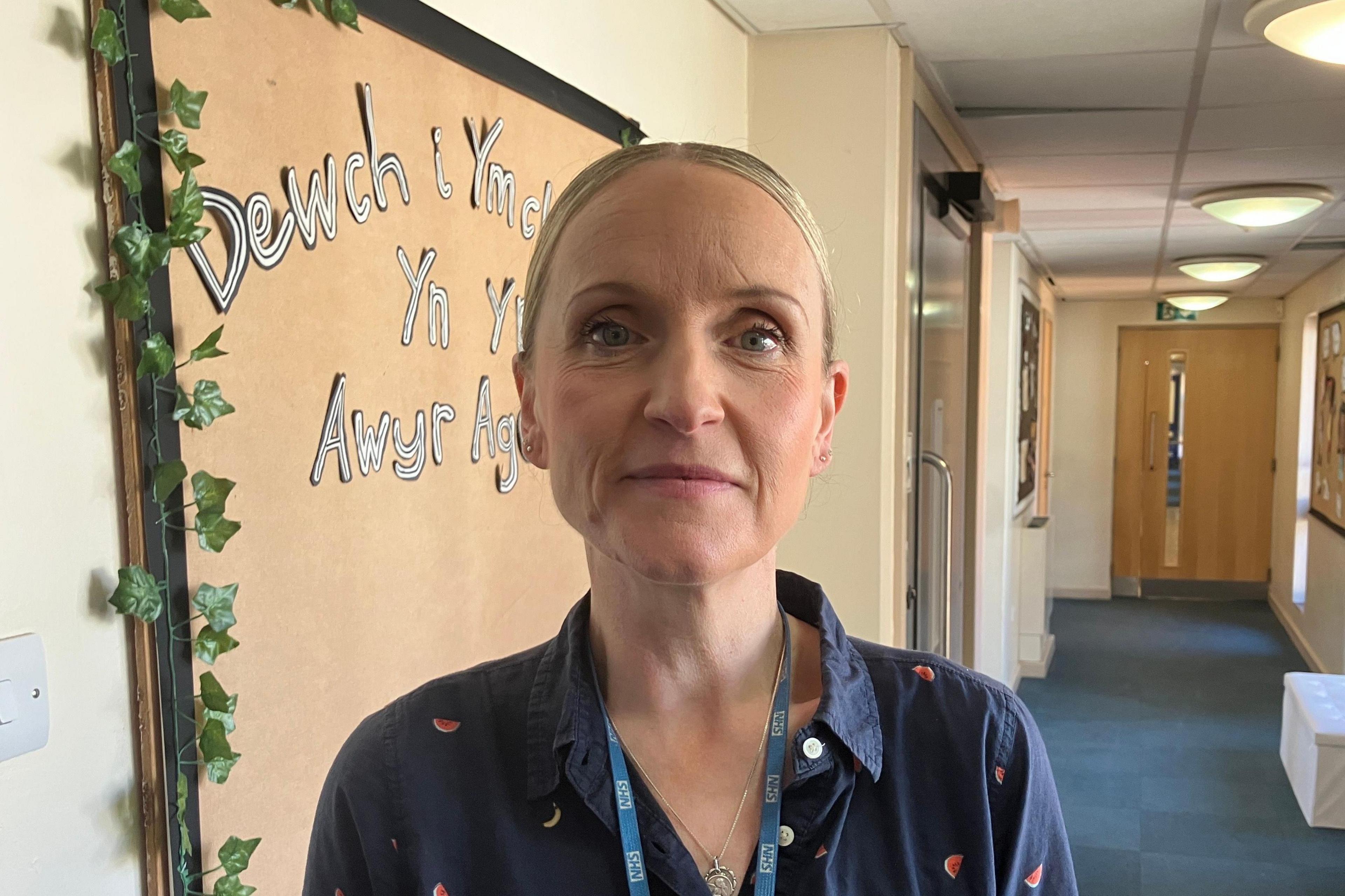 A woman with pulled back blonde hair wearing a blue shirt with a watermelon slice pattern, stands in a school corridor. She is wearing a lanyard with an NHS logo ribbon.