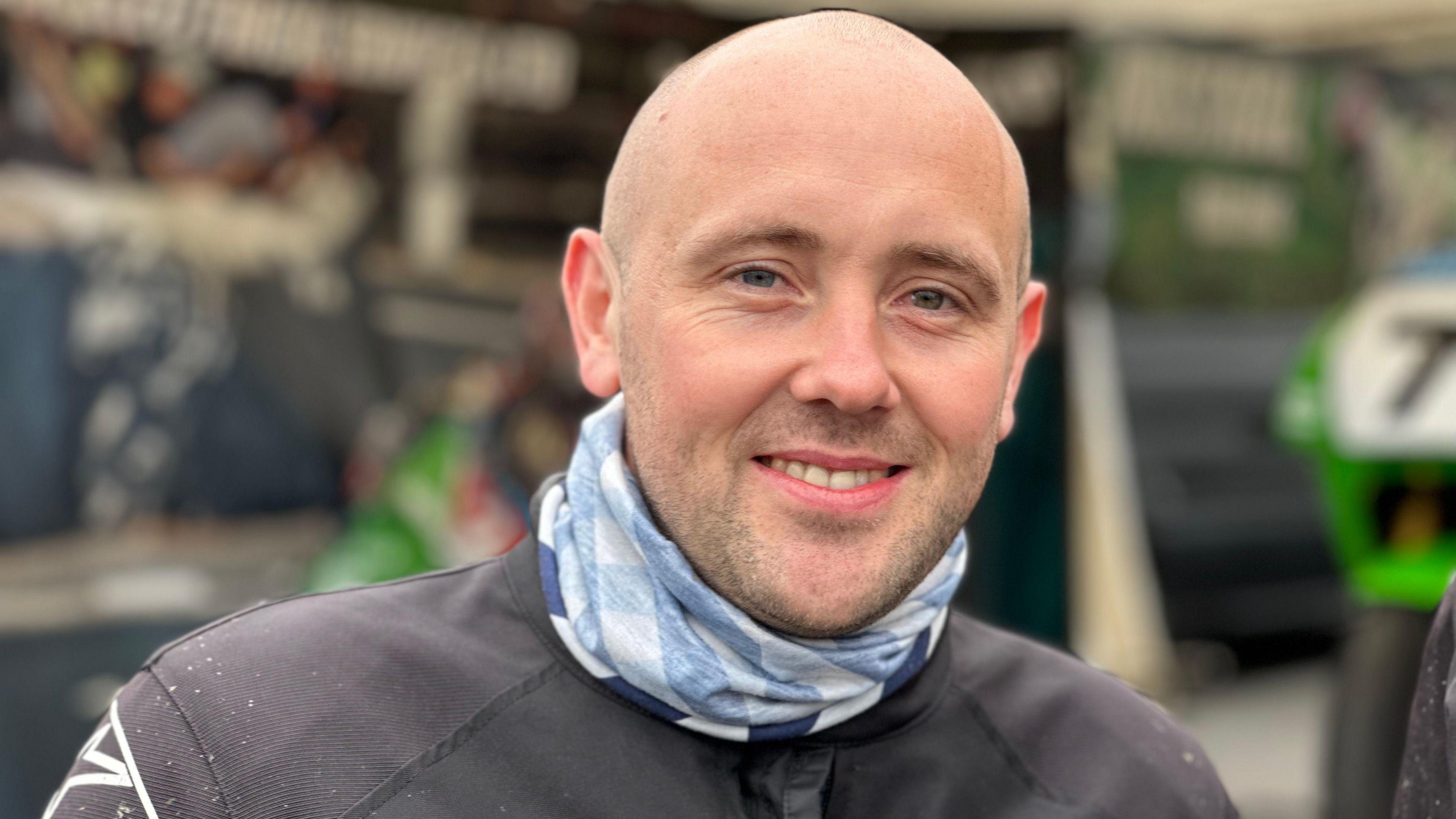 Mark Scott is bald, he has a short beard. He wears a black jacket and blue scarf.