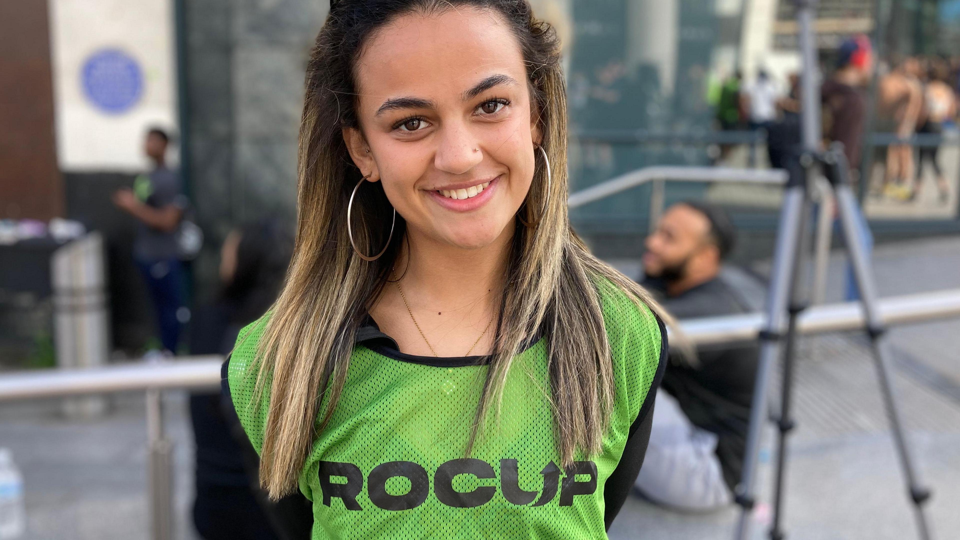 Heniya Azzerghni, who is standing in front of railings in Birmingham city centre. She is smiling at the camera and has sunglasses on her head with long dark hair with blond streaks and large hoop earrings