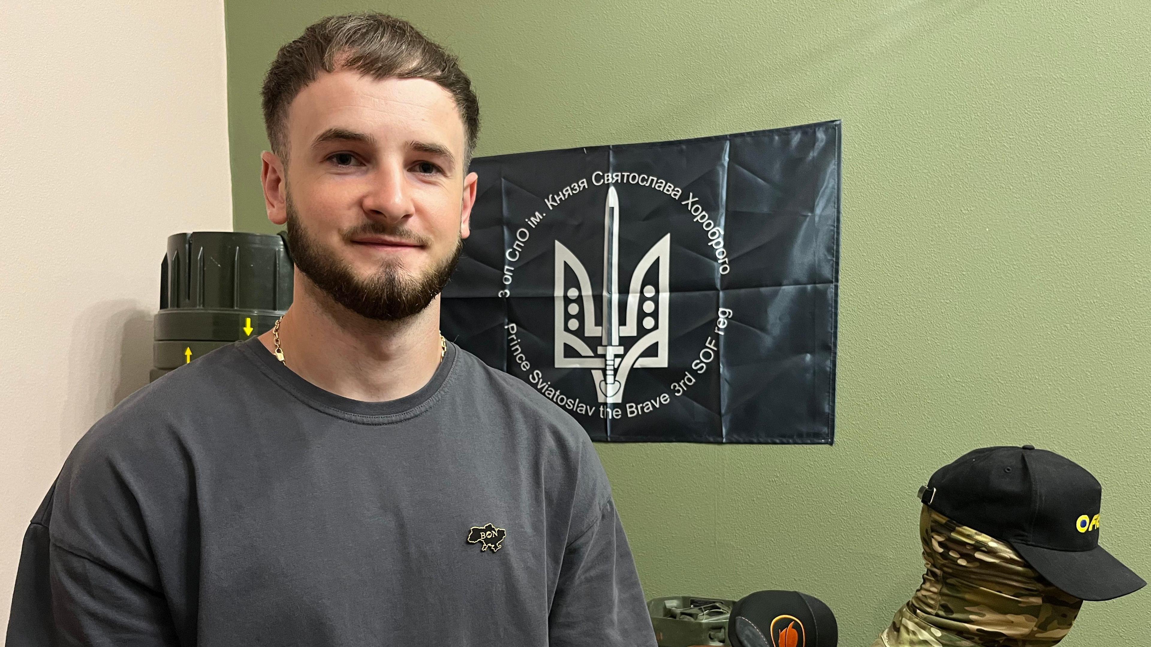 Denys stands with a beard and a grey t-shirt in front of a regimental banner