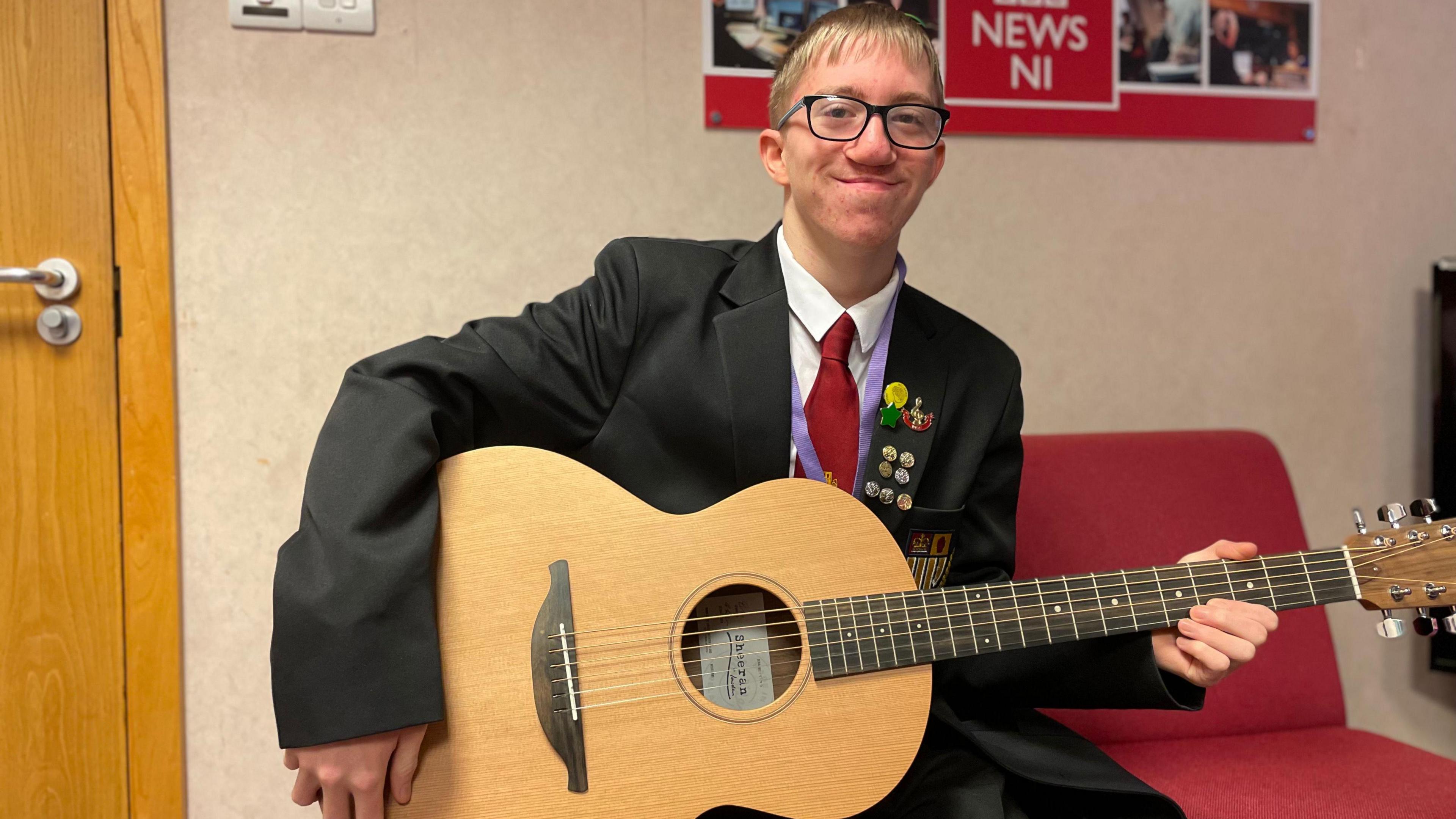 Deacon, a fair-haired teenage boy who wears glasses, holds an hour-glass shaped concert guitar and smiles at the camera. The lapel of his black school blazer, which he wears over a white shirt and maroon tie, is bedecked with badges and pins.
