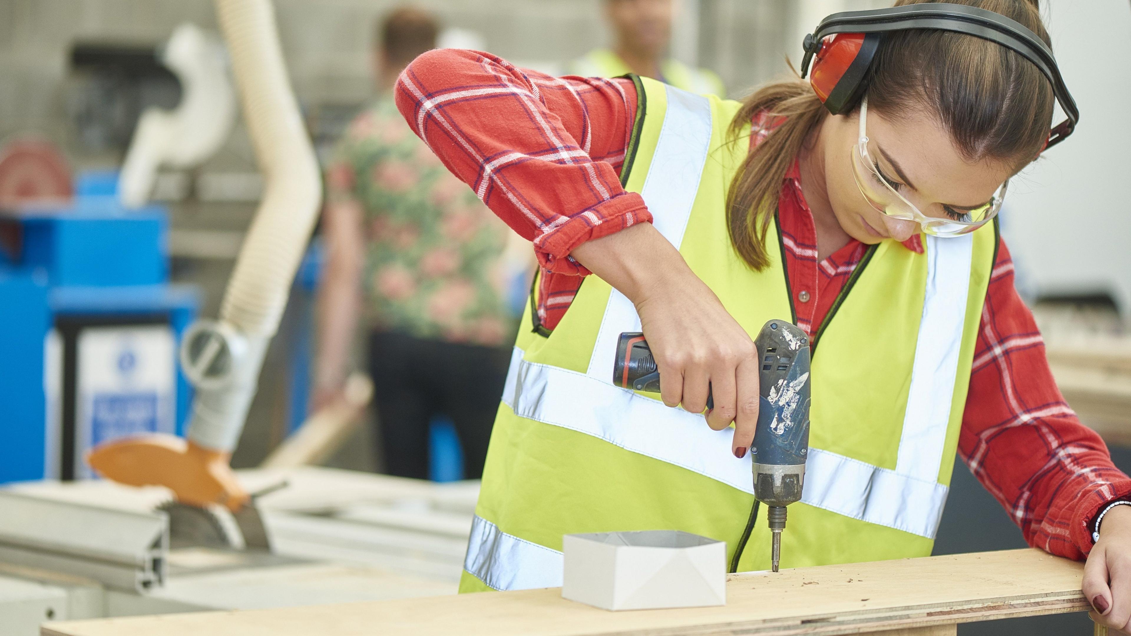 File image of a young woman working in a woodworking workshop while wearing a high-vis jacket and PPE