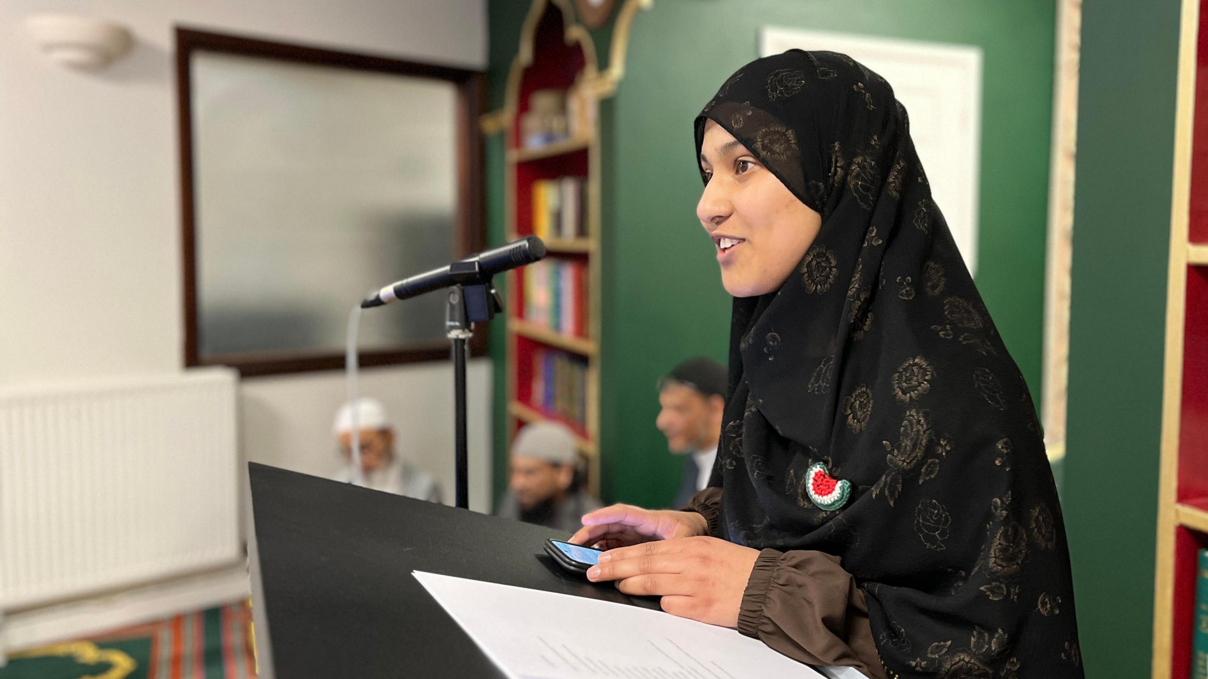 A women in traditional Muslim dress and head covering stands at a lectern at the front of a mosque