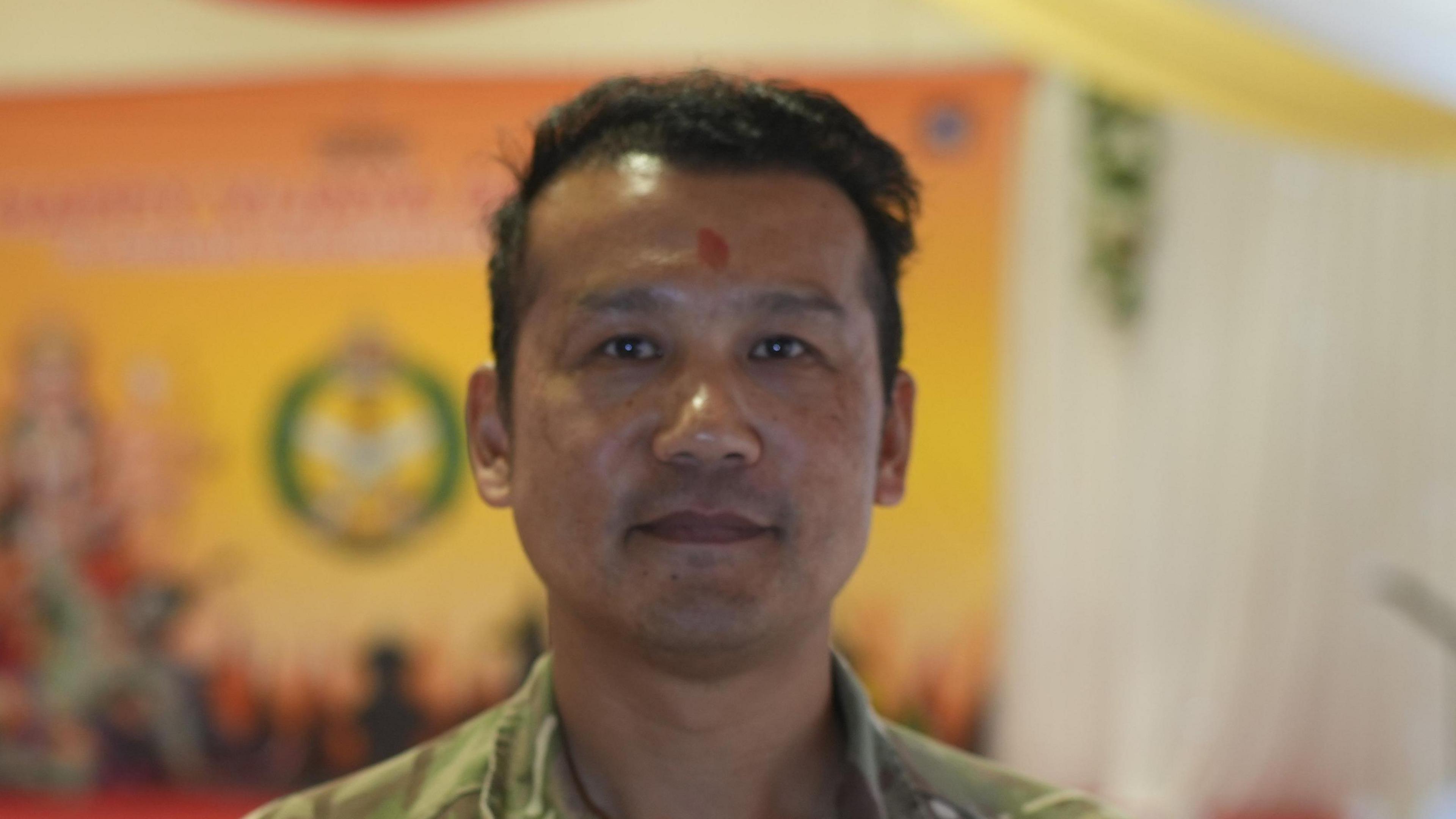 Sgt Maj Nissan Lama smiles at the camera. He has dark hair and wears a green Army uniform. 