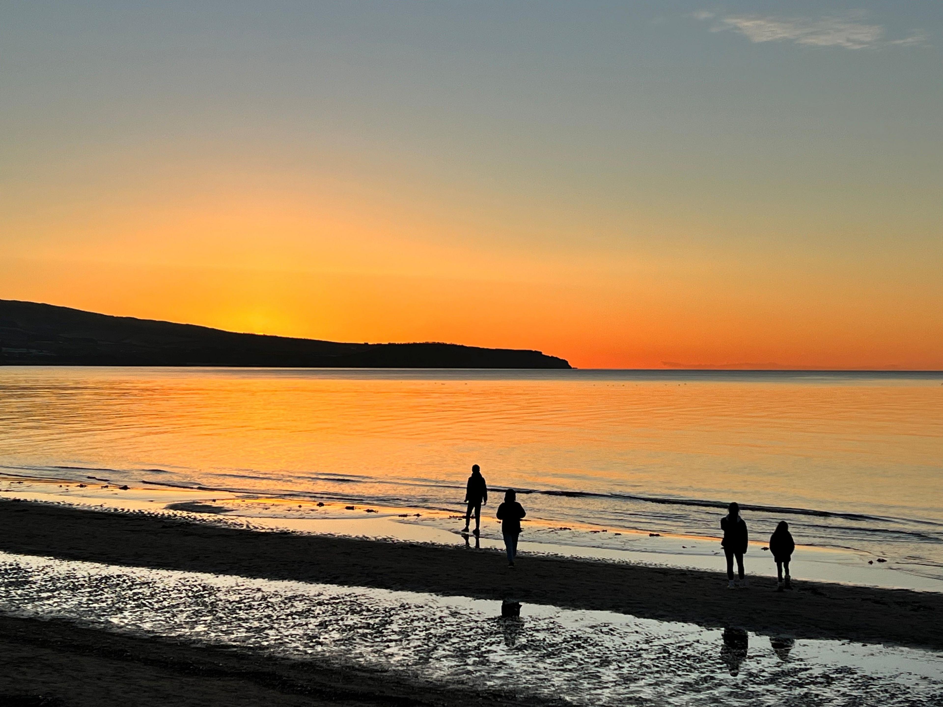 Orange light from a sunset shines down on a beach and the sea, as four silhouetted figures walk along it 