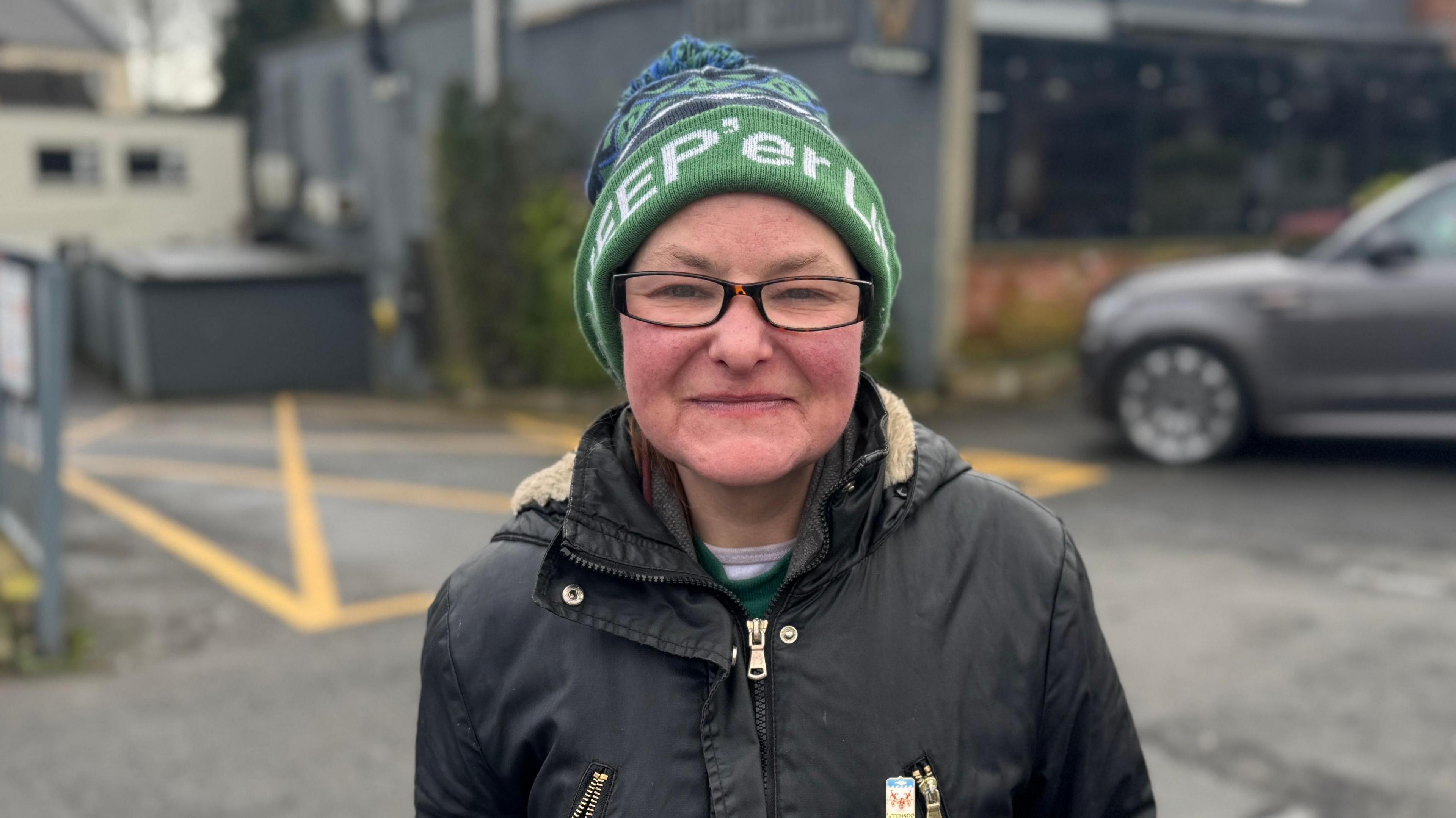 Karla Sweetlove. A woman smiles at the camera while wearing small, rectangular glasses and a green bobble hat with 'Keep'er lit' in white writing. She is wearing a black, zip-up raincoat. She is standing in a car park with a grey building behind her, which is blurred.