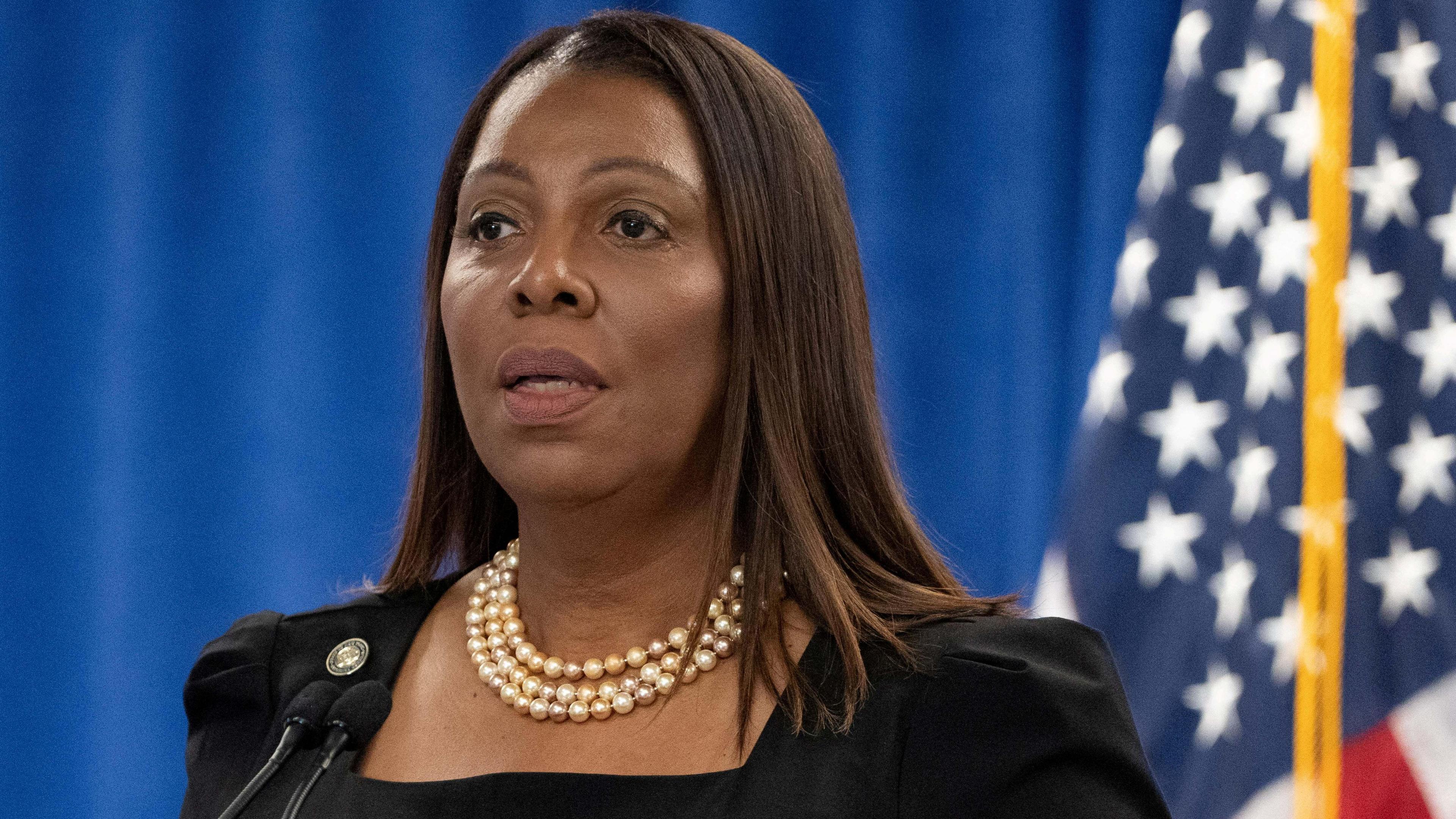 A woman wearing a pearl necklace stands in front of an American flag.