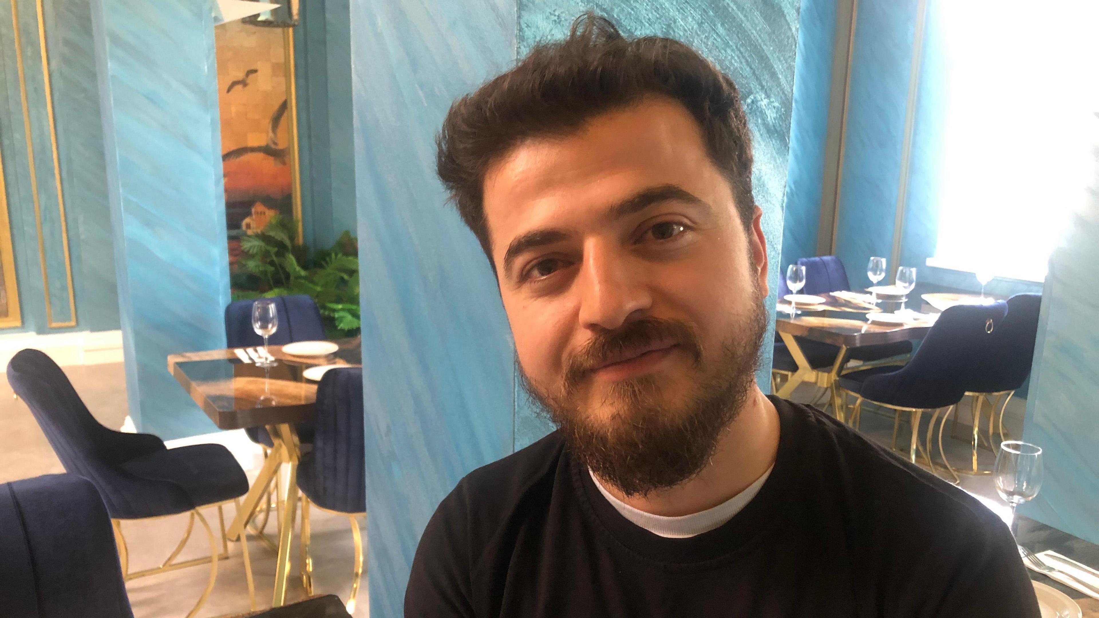 Salih Solak, 28, wearing a black round-collared jumper with white T-shirt underneath. He has dark hair with short beard and moustache. Behind him are blue plush chairs and tables with glasses, plates and cutlery, the restaurant has sky blue walls with blue swirls and a picture is hanging showing birds in flight.