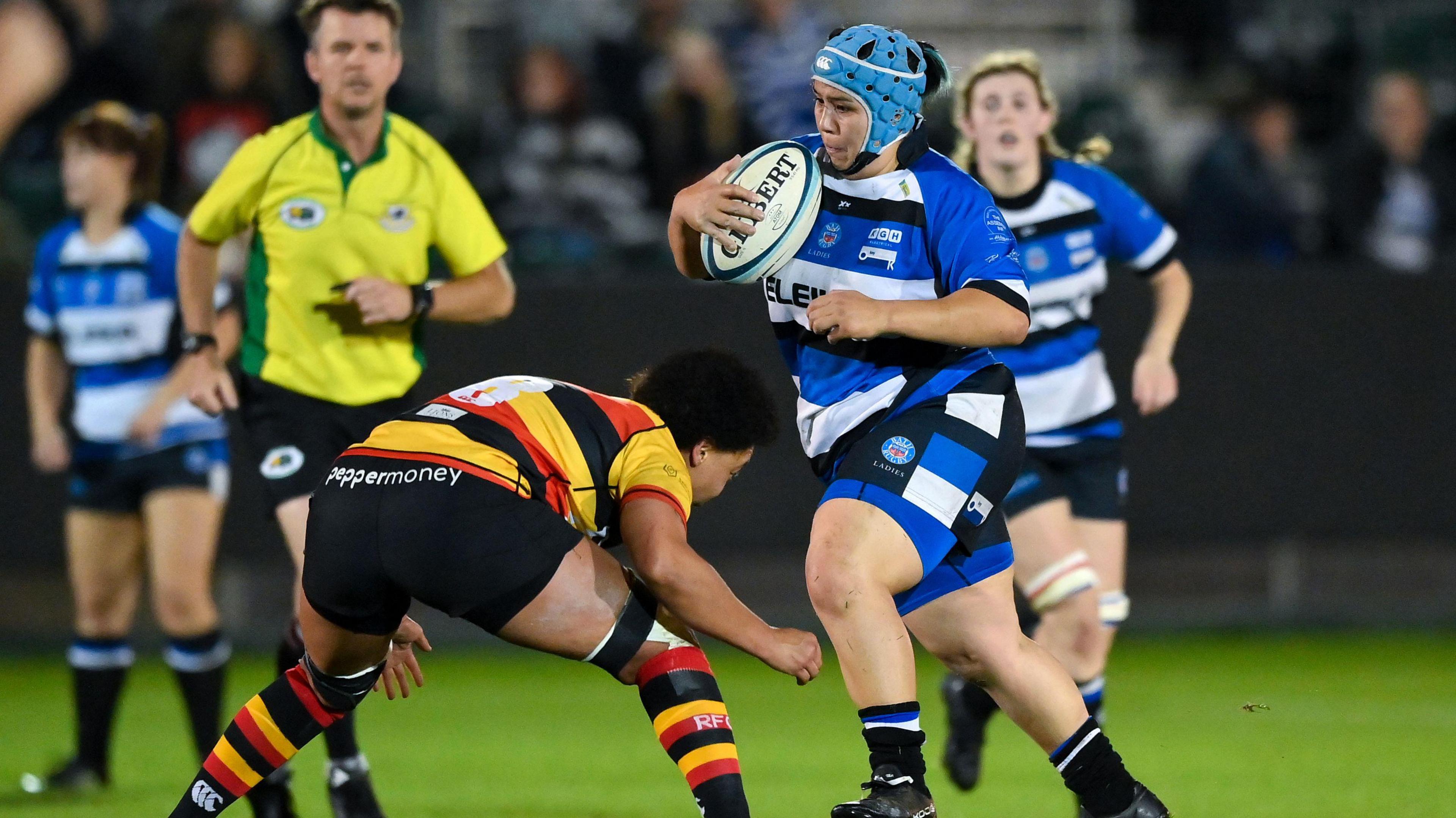 Trish Brown playing a rugby match. She is in a blue and white Bath Ladies outfit with a blue helmet. She is holding the ball and is about to be tackled by a player from the other team.