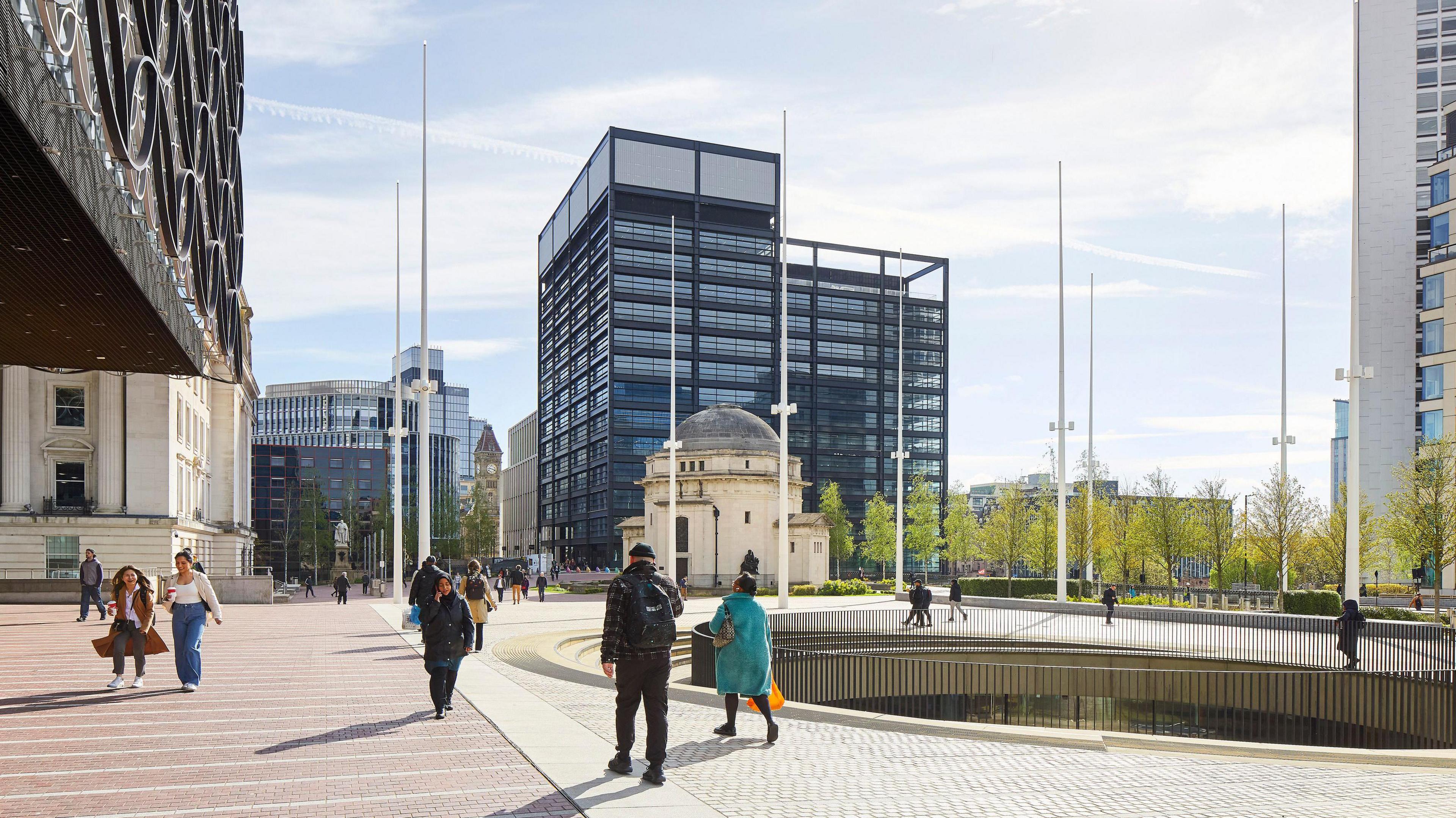 People walking along a paved area in the middle of a city. There are numerous buildings surrounding them, including a tall, black, rectangular structure.