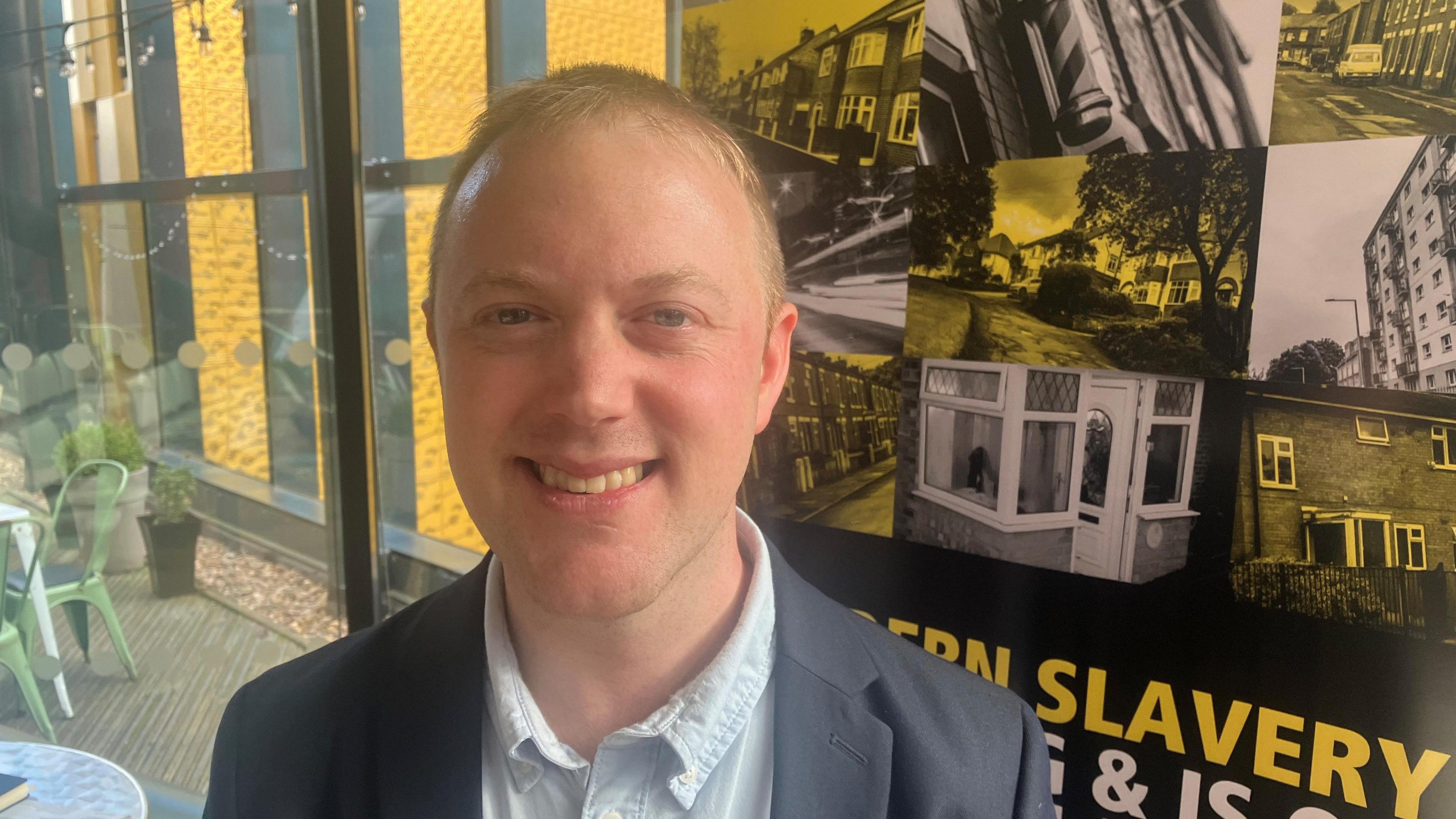 Photograph of Dr Jon Davies - a criminologist at the University of Manchester. He is smiling into the camera, has light short hair and is wearing a light blue short and black blazer. A modern slavery poster is in the background behind him.