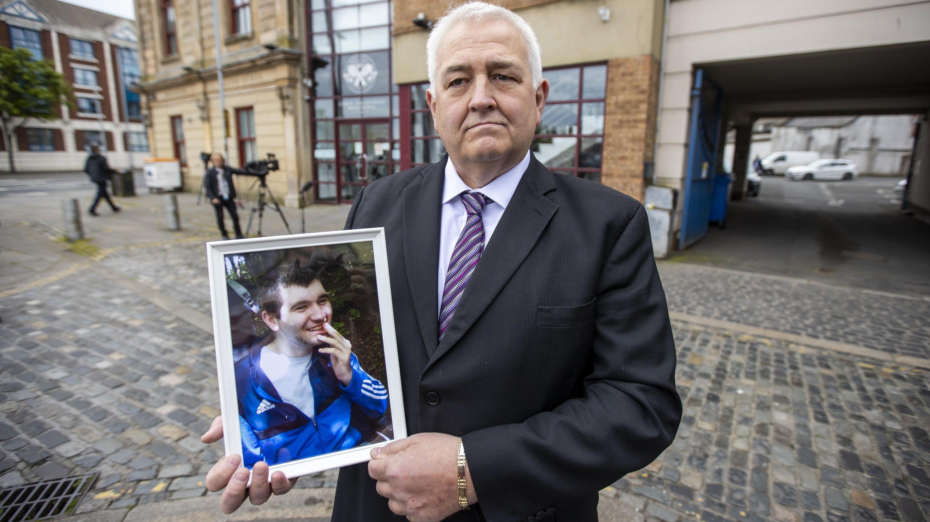 Glynn Brown has short grey hair and is wearing a suit with a striped tie. He is holding a framed photograph of his son Aaron who has black hair and is wearing a blue tracksuit sop and a white tee-shirt