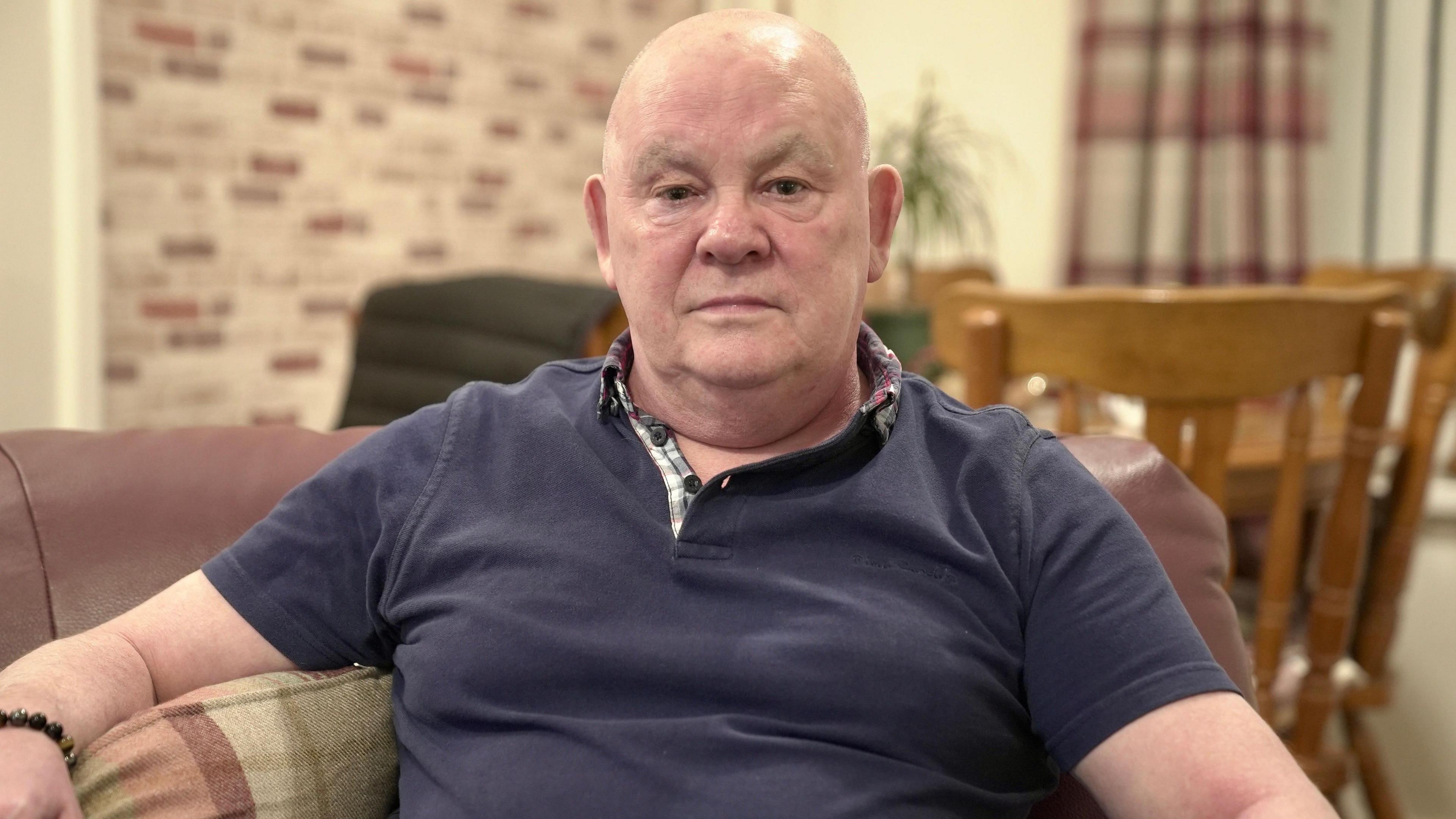 Ralph Morgan is sitting in his living room wearing a navy polo T-shirt, looking straight ahead at the camera. Behind him is a dining table and chairs and a house plant